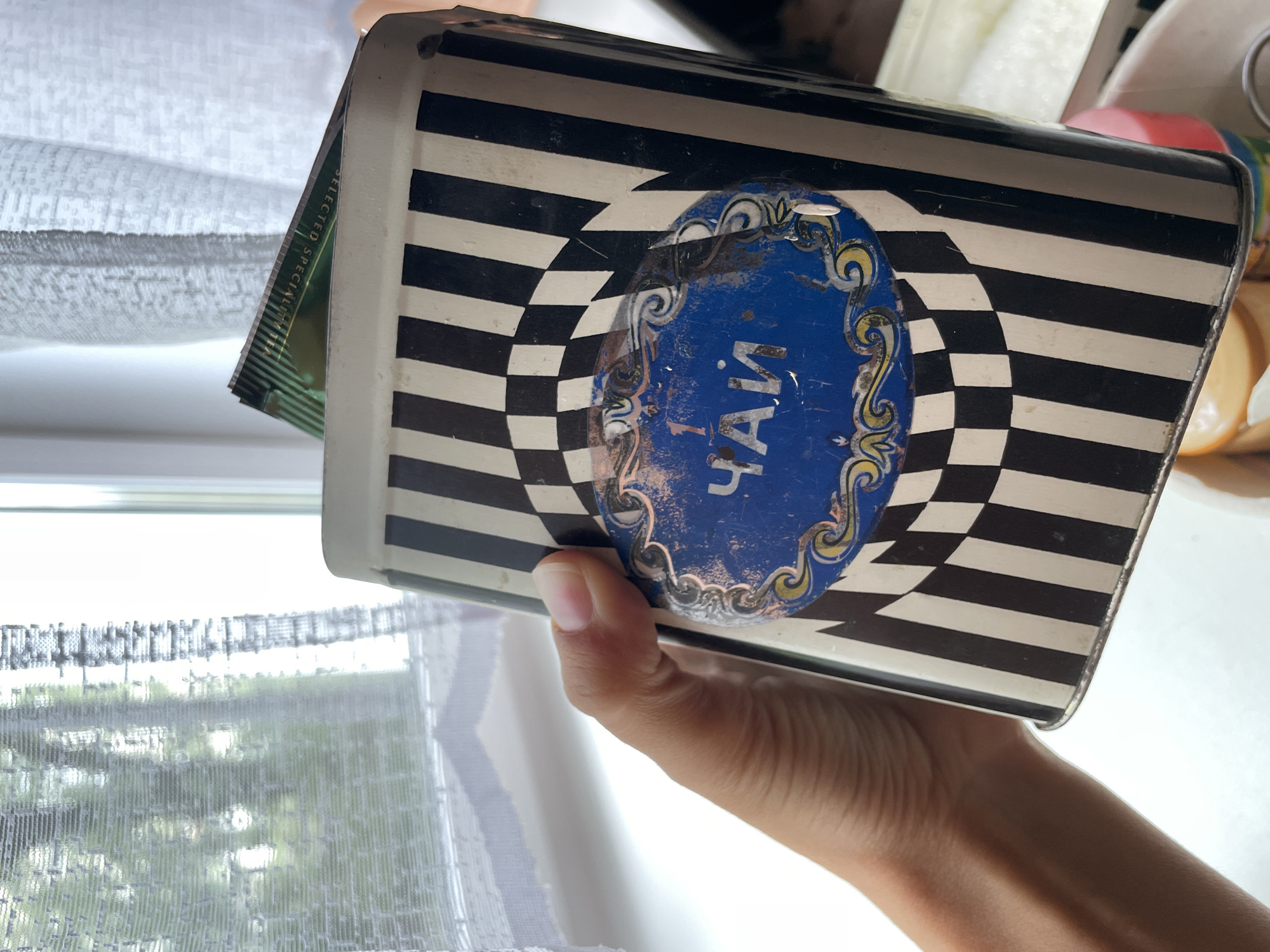 A person is holding a tea caddy. The tea caddy has a checkered pattern and a blue oval with the word "Чай" (tea) written in Russian. The photo appears to have been taken inside near a window with a curtain. There is no information about Zheleznogorsk, Russia.