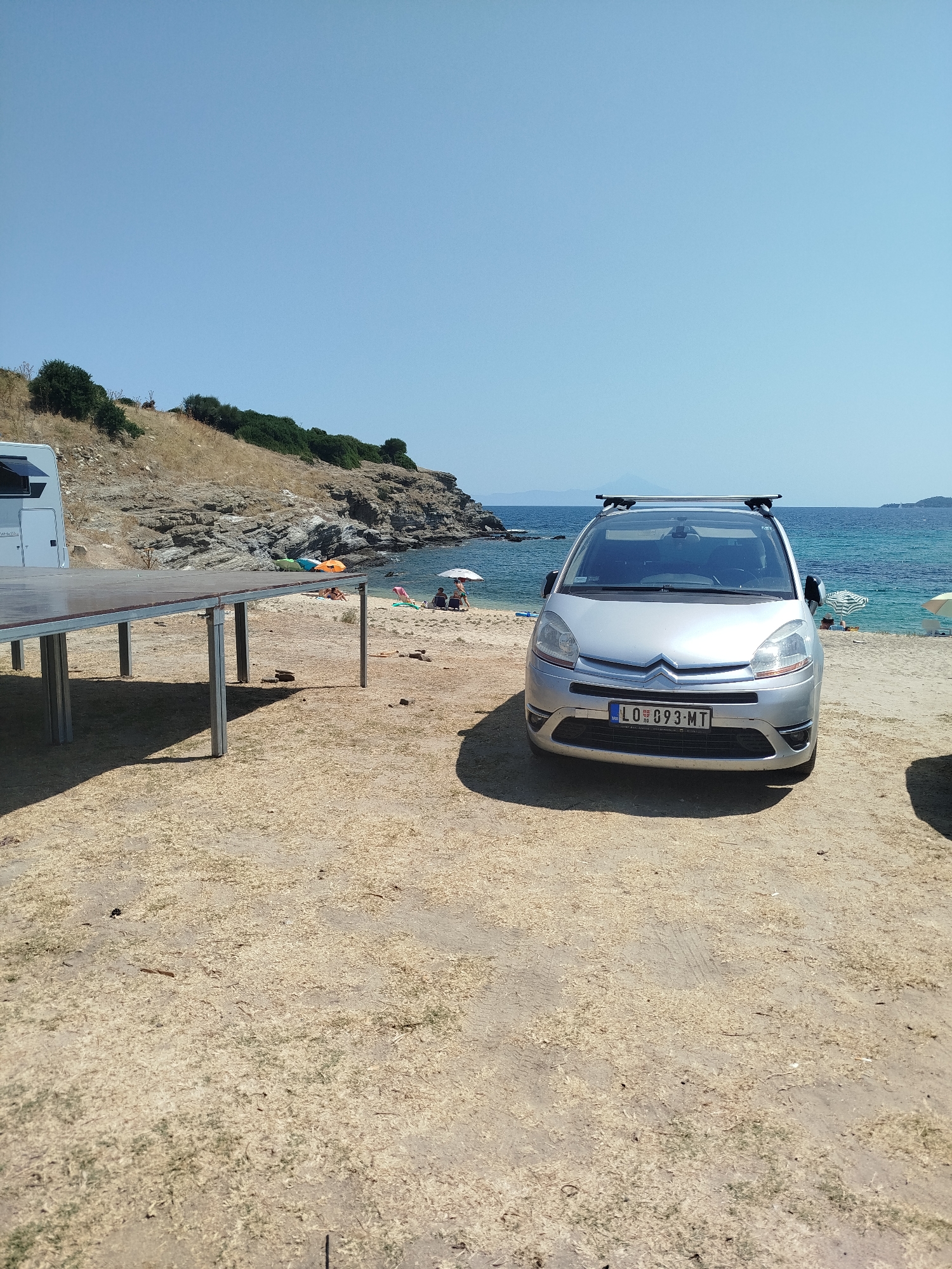 The image depicts a scene on a beach, likely in the Sithonia Municipal Unit, Greece, under a clear blue sky. In the foreground, a silver Citroën C4 Picasso is parked on a sandy and dry grass-covered area. The car has a roof rack. To the left of the car, there is a raised wooden platform supported by metal legs. Further back, along the coastline, is a rocky, vegetated hill. A section of the sea is visible, with a few people relaxing on beach towels and umbrellas. The overall environment suggests a sunny day at a coastal location, possibly a campsite or a public beach with amenities.
