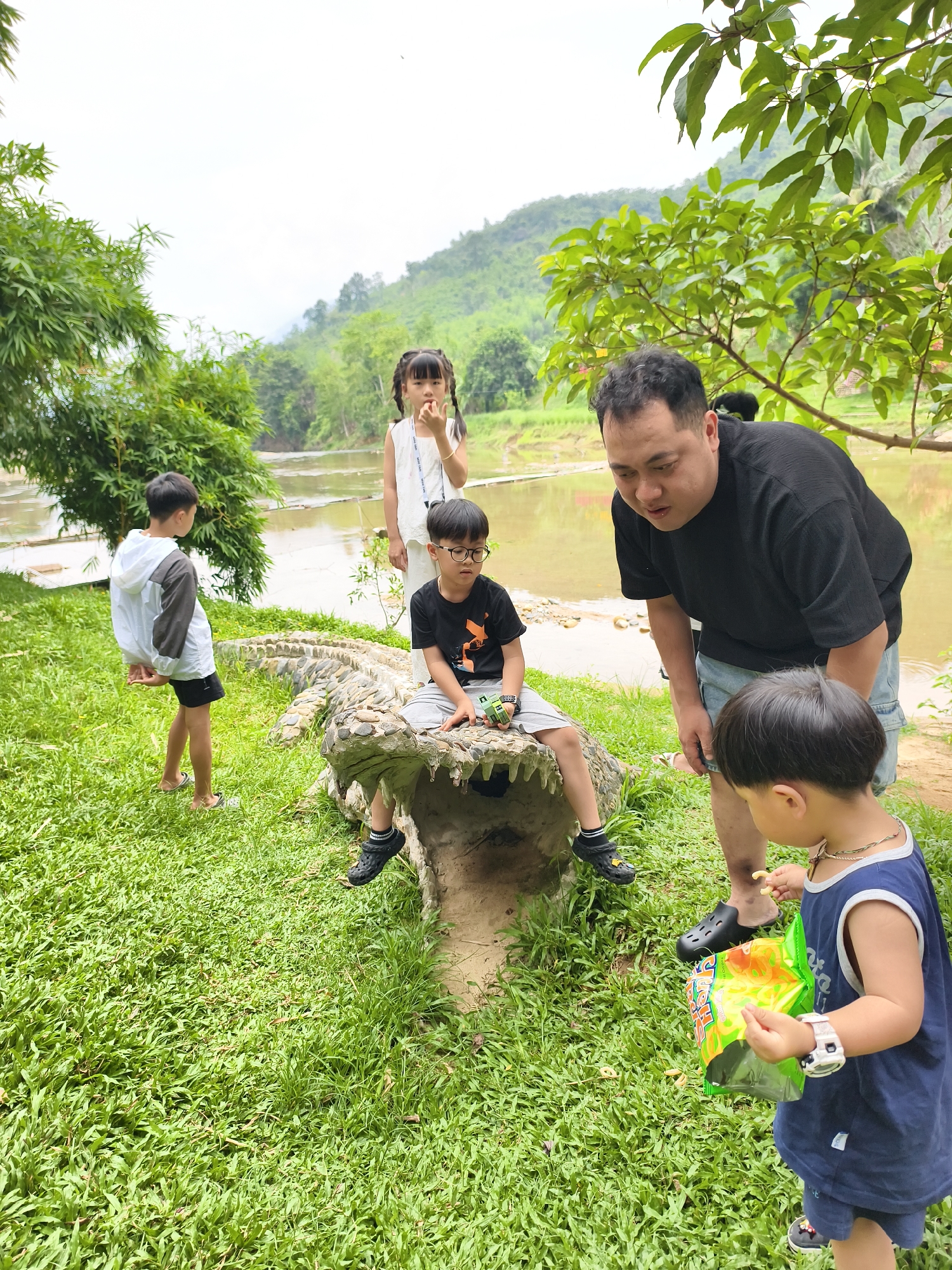 In Nam Khanh Vinh, Vietnam, a group of children and an adult man are gathered around a concrete alligator statue near a body of water. One child sits on the statue's open mouth, wearing glasses, a black t-shirt, and grey shorts. Another child, wearing a white dress, stands beside the alligator statue. A third child wearing a white jacket stands to the left of the statue and looks away. A younger child, wearing a blue tank top and shorts, holds a bag of chips while looking at the adult male, who wears a black t-shirt and denim shorts, leaning over the boy with the chips. The environment consists of a grassy area and a tree line with a mountain range in the background.