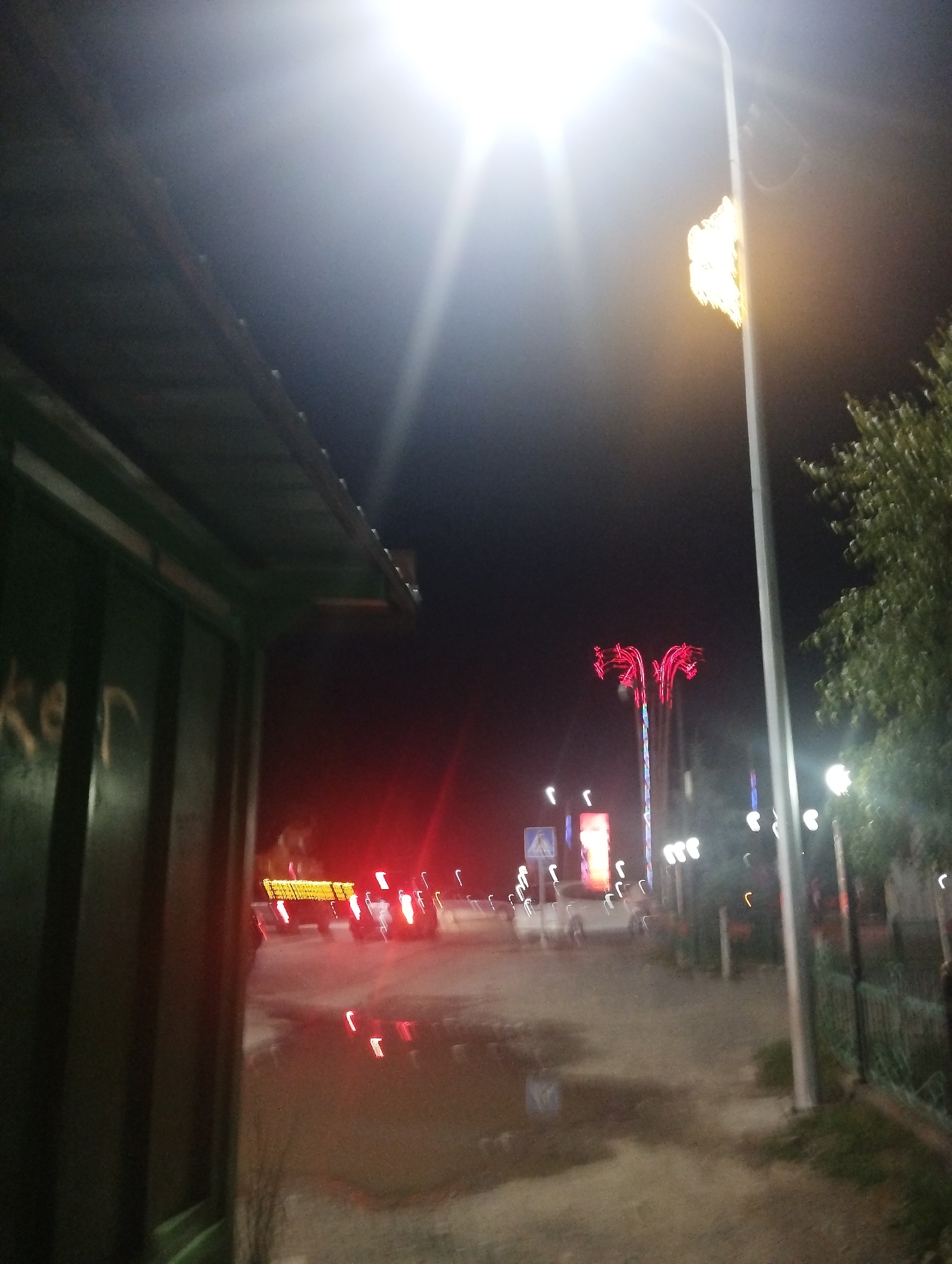 The image captures a night scene on a street in Naryn City, Kyrgyzstan. A bright overhead light illuminates the foreground, casting a strong glare. To the left, the side of a dark green, metallic structure, possibly a container or building facade, is visible with faint white markings. In the mid-ground, a street stretches into the distance, featuring various light sources including streetlights, decorative red lights resembling palm trees, and illuminated signs. A white vehicle is parked on the right side of the street, and further down, what appears to be a truck with illuminated yellow lights is driving away, its taillights visible. The ground surface is uneven and appears wet in places, reflecting the surrounding lights. A tree with green foliage is present on the right side of the frame. The overall impression is a somewhat blurry, low-light urban environment at night.