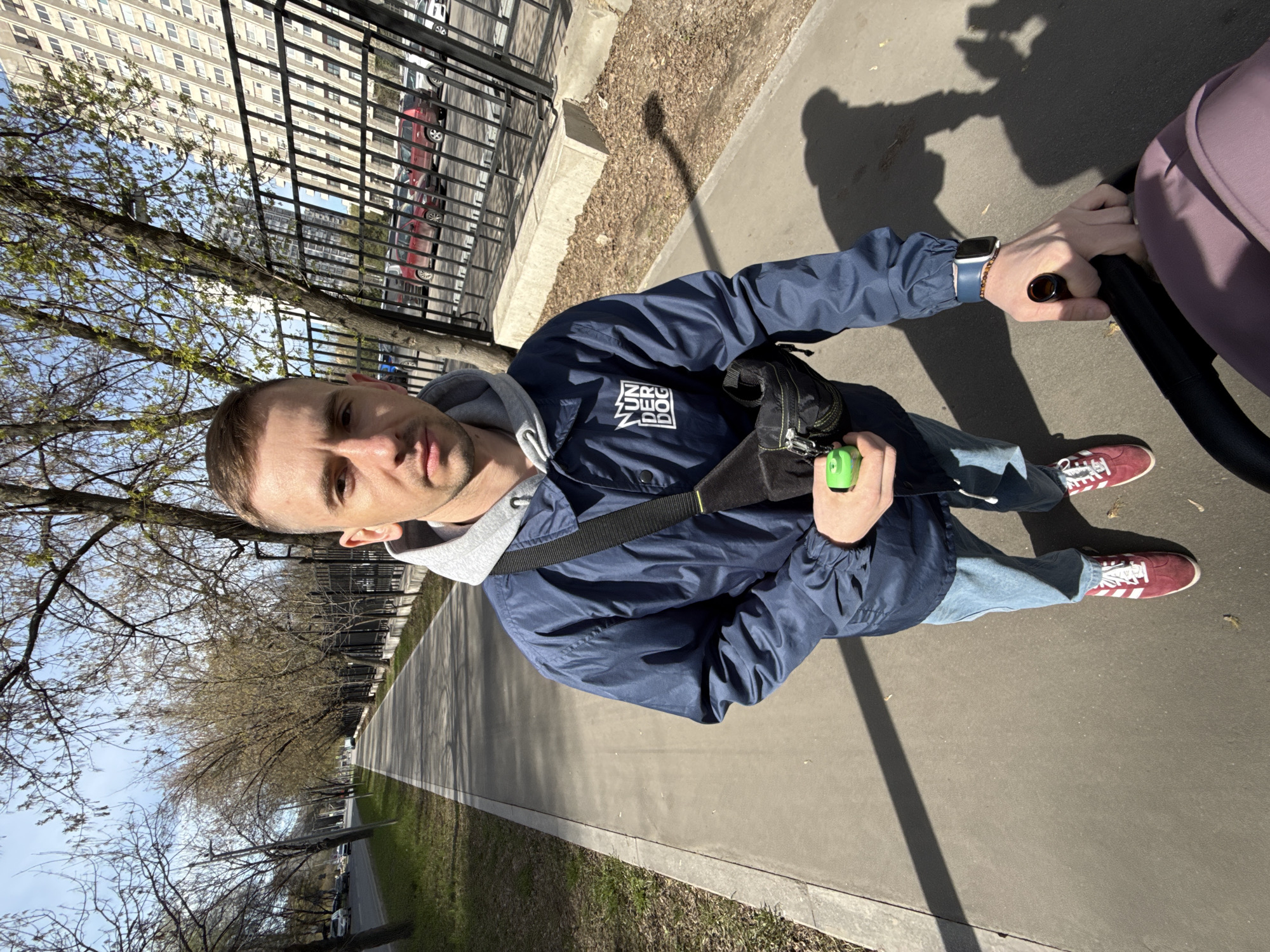 A man in his 20s or 30s is captured in a high-angle shot, looking directly at the camera while pushing a baby stroller outdoors on a sunny day. He is dressed in a navy blue bomber jacket over a grey hooded sweatshirt, light blue jeans, and maroon Adidas sneakers with white stripes. A black fanny pack or crossbody bag is slung across his chest. On his left wrist, he wears an Apple Watch with a blue band. His right hand holds a small green bottle, while his left hand grips the handle of a light pink/lavender baby stroller, partially visible on the right side of the frame.
The setting appears to be a paved path or sidewalk in an urban or suburban park-like area. Strong shadows of the man, the stroller, and a lamppost are cast on the concrete, indicating bright sunshine, likely mid-morning or afternoon. The sky is clear blue. Along the path, there are numerous trees, mostly bare or just beginning to bud, suggesting early spring. A black metal fence runs parallel to the path, behind which tall, light-colored apartment buildings with many windows are visible. Several cars are parked along a street adjacent to the fence. The man's expression is neutral.
On the front of his navy jacket, there is a stylized white logo with text that appears to read "MUR" stacked above "NEO", which is then stacked above "DOG".