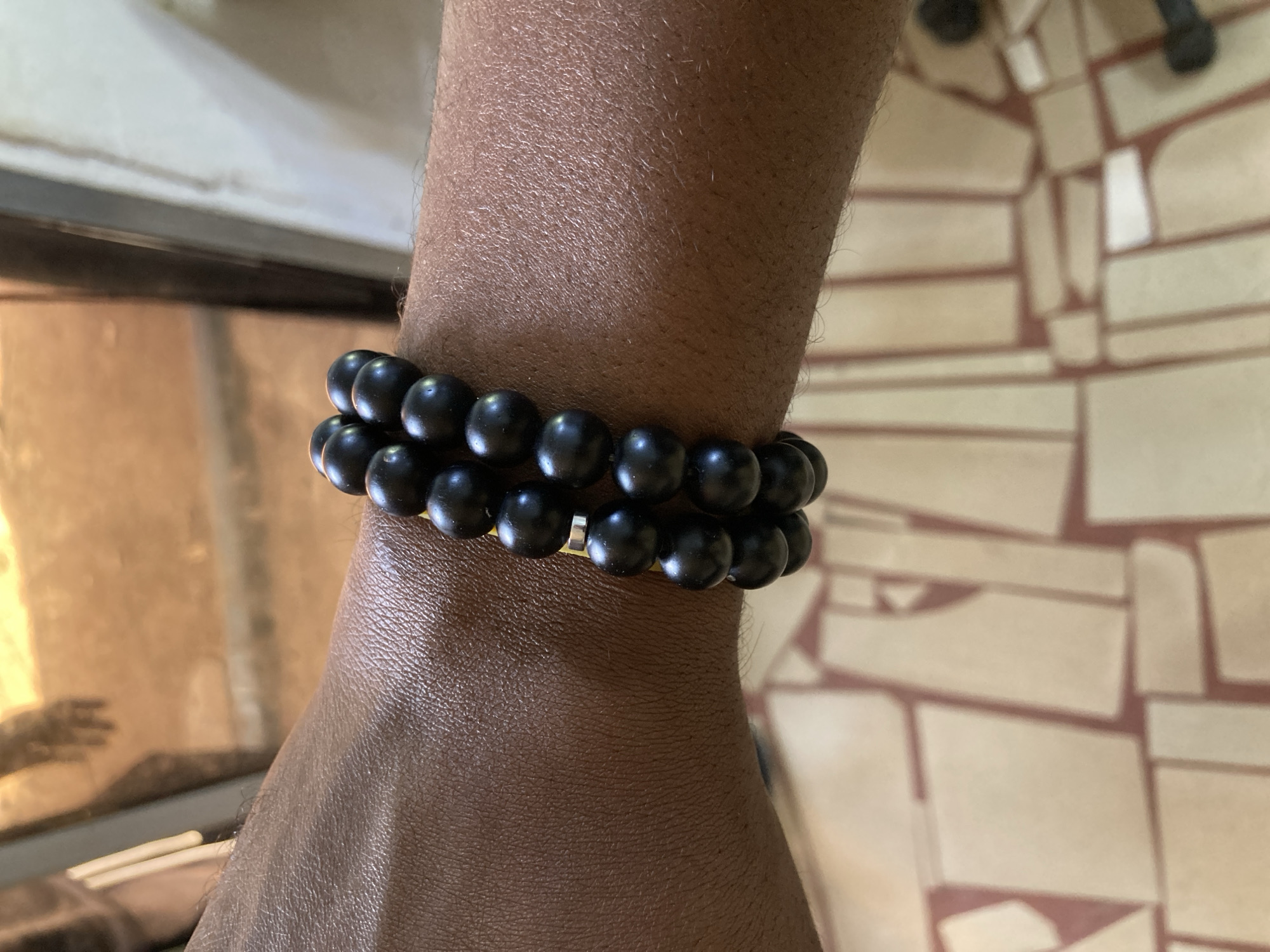The image shows a close-up of a brown-skinned wrist wearing two strands of black beaded bracelets. The background is a blurred mix of light-colored tile patterns and a reflective surface that suggests glass or a polished surface. There's no information to determine the city or country in the image.