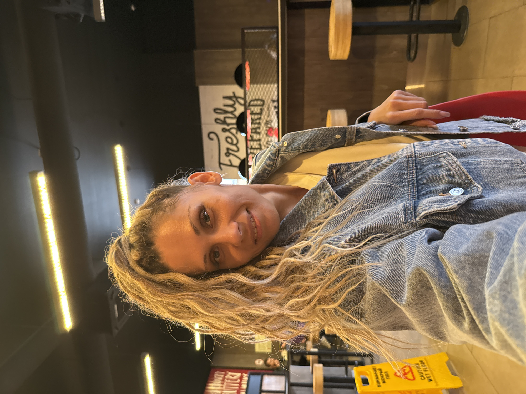 The image shows a selfie of a woman with blonde, curly hair pulled into a high ponytail. She is wearing a light yellow t-shirt and a blue denim jacket. Her facial expression is a slight smile, and she is looking directly at the camera. In the background, a sign that reads "Freshly Prepared" is visible, suggesting the location is a food establishment. There are also bar stools and lighting fixtures in the background, consistent with a restaurant or cafe setting. A yellow caution sign with a wet floor symbol is partially visible on the left side of the image. The overall environment appears to be indoors, with dark ceilings and illuminated elements.