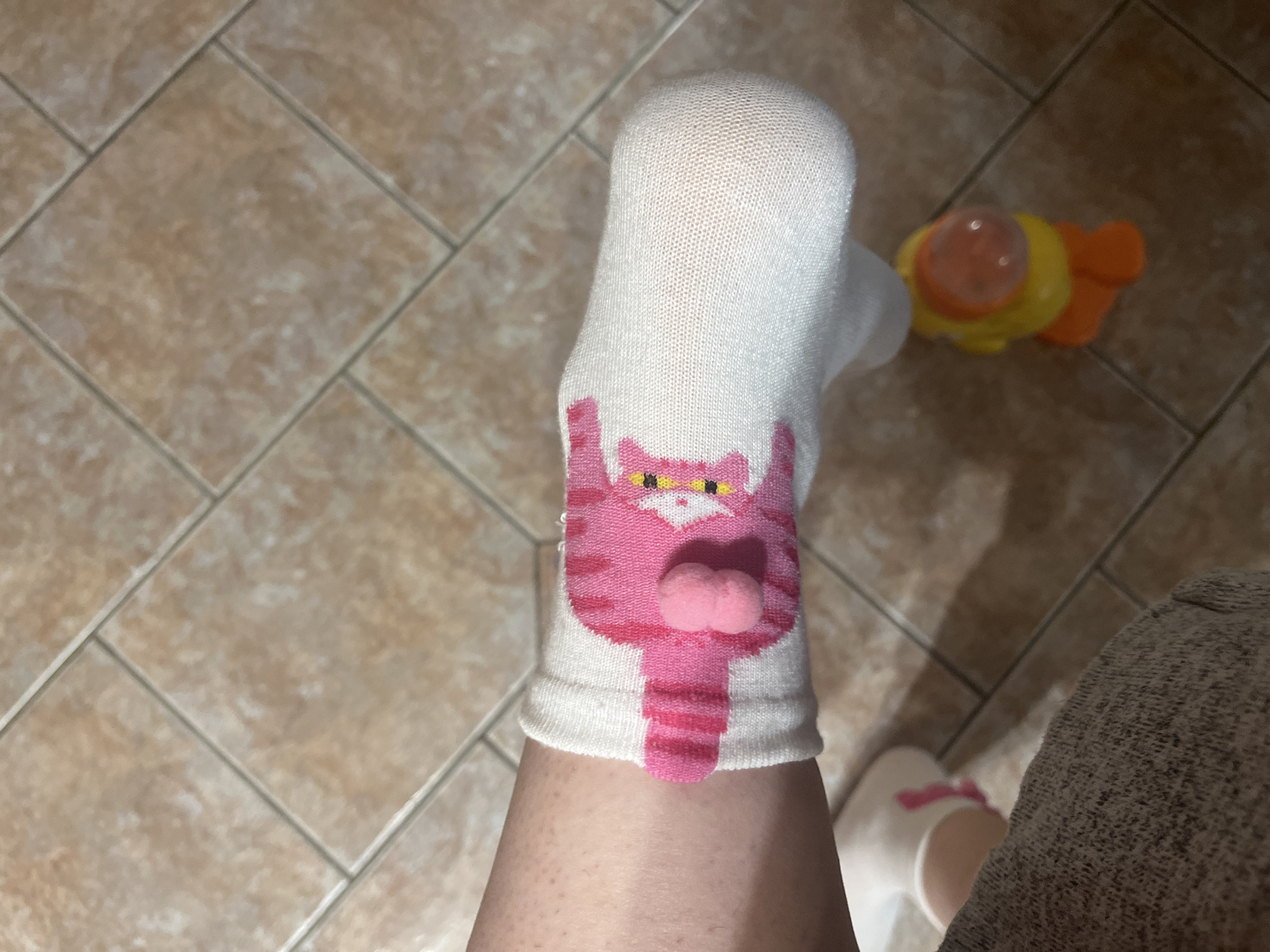 The image depicts a person wearing a white sock with a pink cat design on their foot, which is resting on a tiled floor. A yellow and orange toy is visible nearby. Another foot wearing a white sock with a pink bow is also visible. No location details are available, particularly not Novosibirsk, Russia.