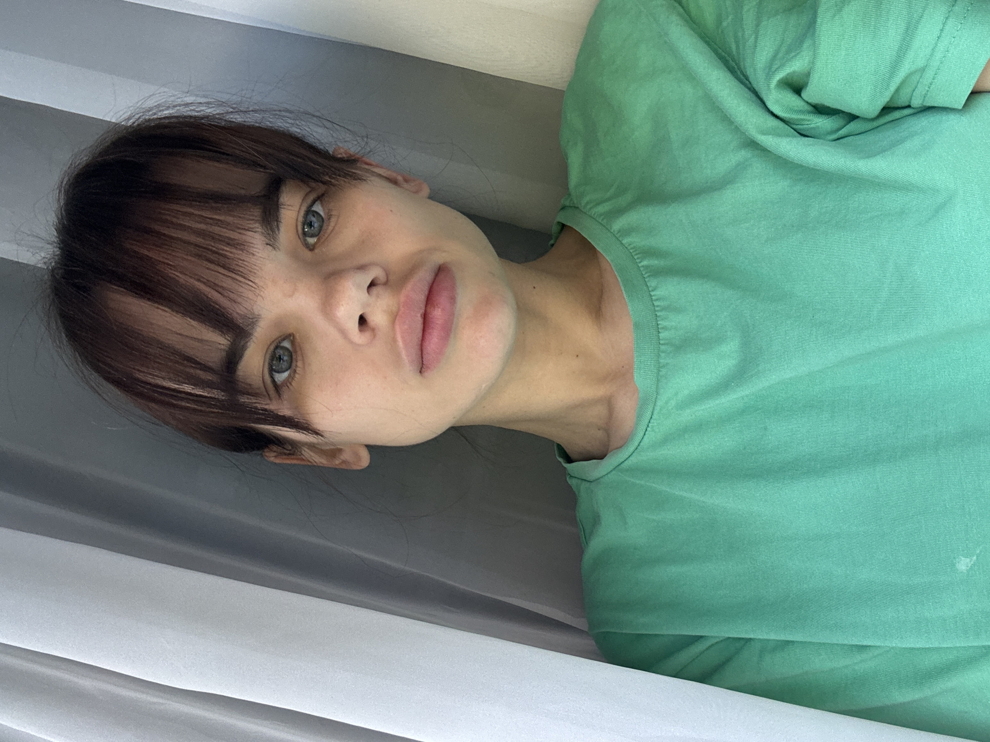 A woman with light skin, brown hair with bangs, and greyish-blue eyes is wearing a green t-shirt. She is looking at the camera. The background appears to be a white and grey striped curtain or wall. There is no indication of the city Rostov-on-Don, Russia.