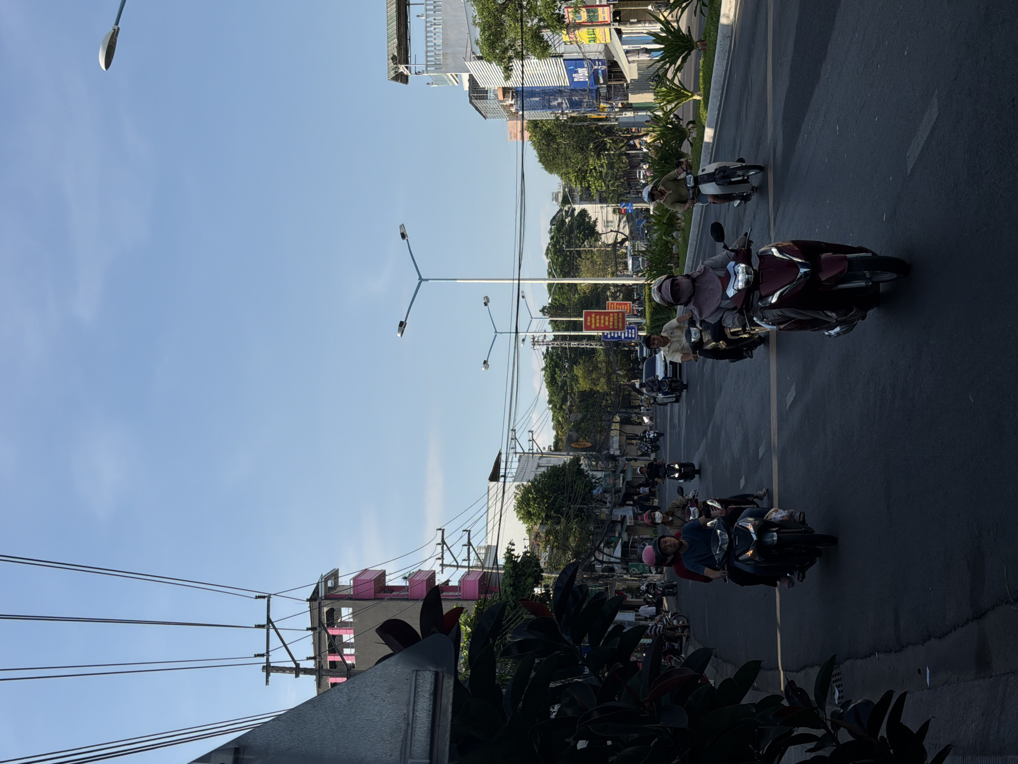 The image depicts a street scene in Bac Nha Trang Ward, Vietnam, under a clear blue sky. The foreground shows a dark, leafy plant on the left, with a section of a gray building structure. The street is paved and appears to be a busy thoroughfare with multiple lanes. Several individuals are riding motorcycles in various directions. In the immediate foreground, a dark maroon scooter with a person wearing a light pink headscarf and a patterned mask is being ridden. To the left of this, another scooter carries two individuals; the rider wears a pink helmet, and the passenger behind has a dark helmet and a light-colored shirt. Further down the street, other motorcyclists are visible, some individually and some in pairs, with a mix of helmets and masks being worn by the riders.
The environment features buildings lining both sides of the street. On the left, a pink building with multiple stories and balconies is prominent, with power lines and poles running overhead. On the right, buildings are closer to the street, with some displaying signage, including what appears to be a blue sign for "BV Bank." A median strip with palm-like plants runs down the center of the road in the distance. Streetlights are also visible on tall poles along the street. The overall impression is one of daily urban activity, with motorcycles being the primary mode of transport.