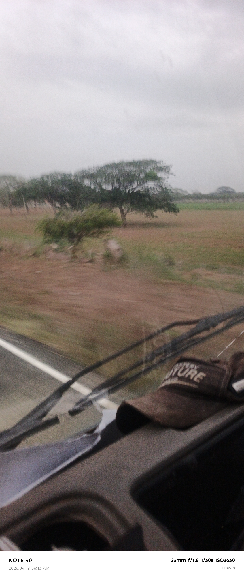 Here is a detailed description of the image:
**Scene**: A photograph taken from inside a moving vehicle, likely a car or bus, showing a rural landscape outside. The image is blurred due to motion.
**Environment**: The landscape consists of a flat, open field with brown vegetation. Several trees are scattered across the field, appearing in the middle and background. The sky is overcast with a greyish tone, suggesting a cloudy or rainy day.
**Objects**: In the foreground, the vehicle's interior is visible. A sun visor with the word "NATURE" on it lies on the dashboard near the windshield. The windshield and wipers can also be seen.
**Textual Information**: The bottom of the image contains text indicating the following:
* "NOTE 40"
* Date and time: "2026.04.19 06:13 AM"
* Camera settings: "23mm f/1.8 1/30s ISO3630"
* Location: "Tinaco"
**Location Details**:
* The camera settings text indicates that the location is "Tinaco." Tinaco is a city in Arismendi Municipality, Cojedes State, Venezuela.