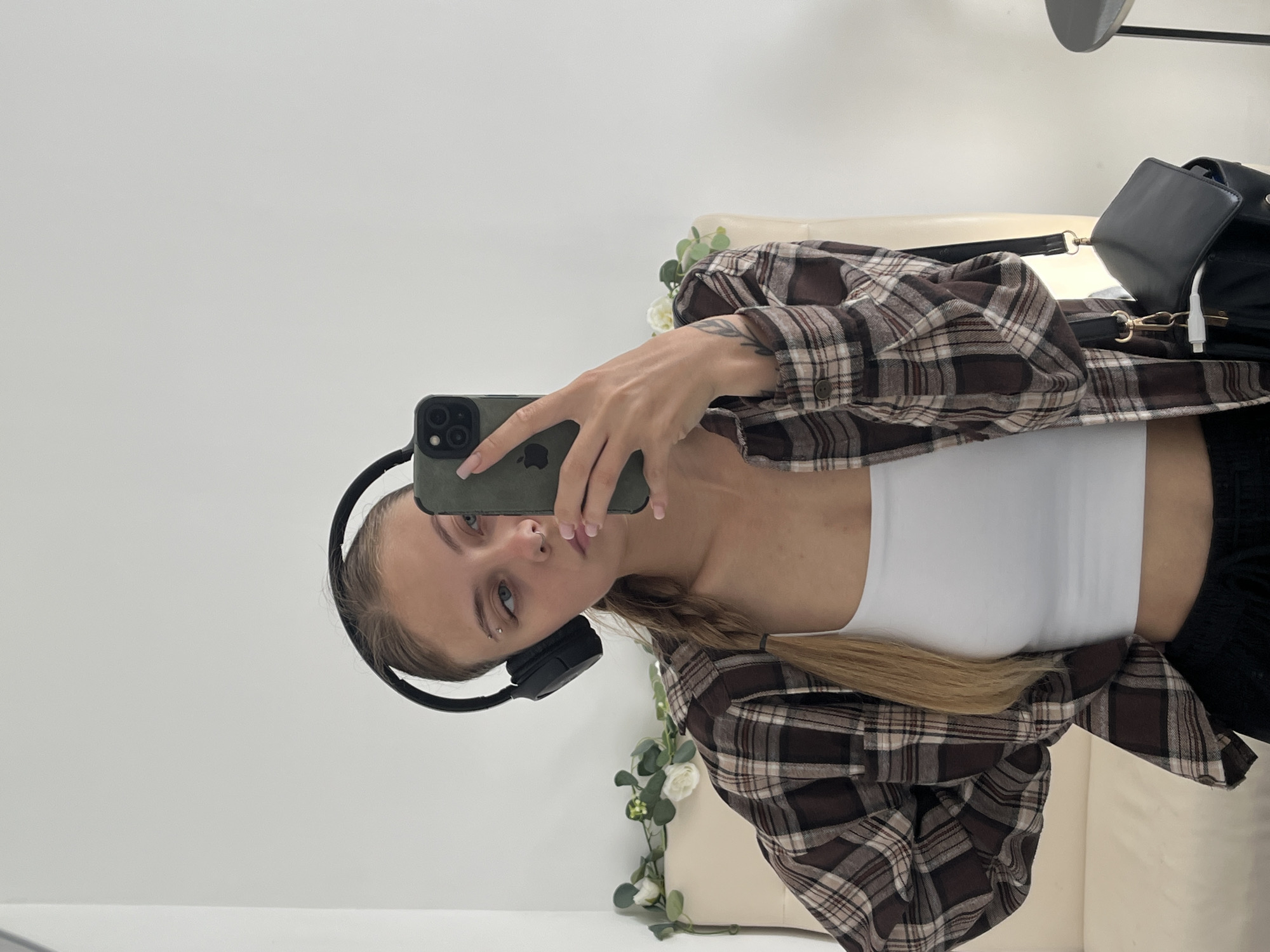 A young Caucasian woman with blonde hair in a braid is taking a mirror selfie indoors. She is wearing headphones, a white tank top, and an open brown and beige plaid shirt. She has a piercing above her eyebrow and a tattoo on her forearm. She is holding a smartphone in her right hand, angled to capture her reflection. A black bag with a white charging cable is slung over her shoulder. The background appears to be a room with a white wall and a light-colored couch adorned with artificial white flowers and green leaves. There is no indication of the city of Фоки, Russia in the image.