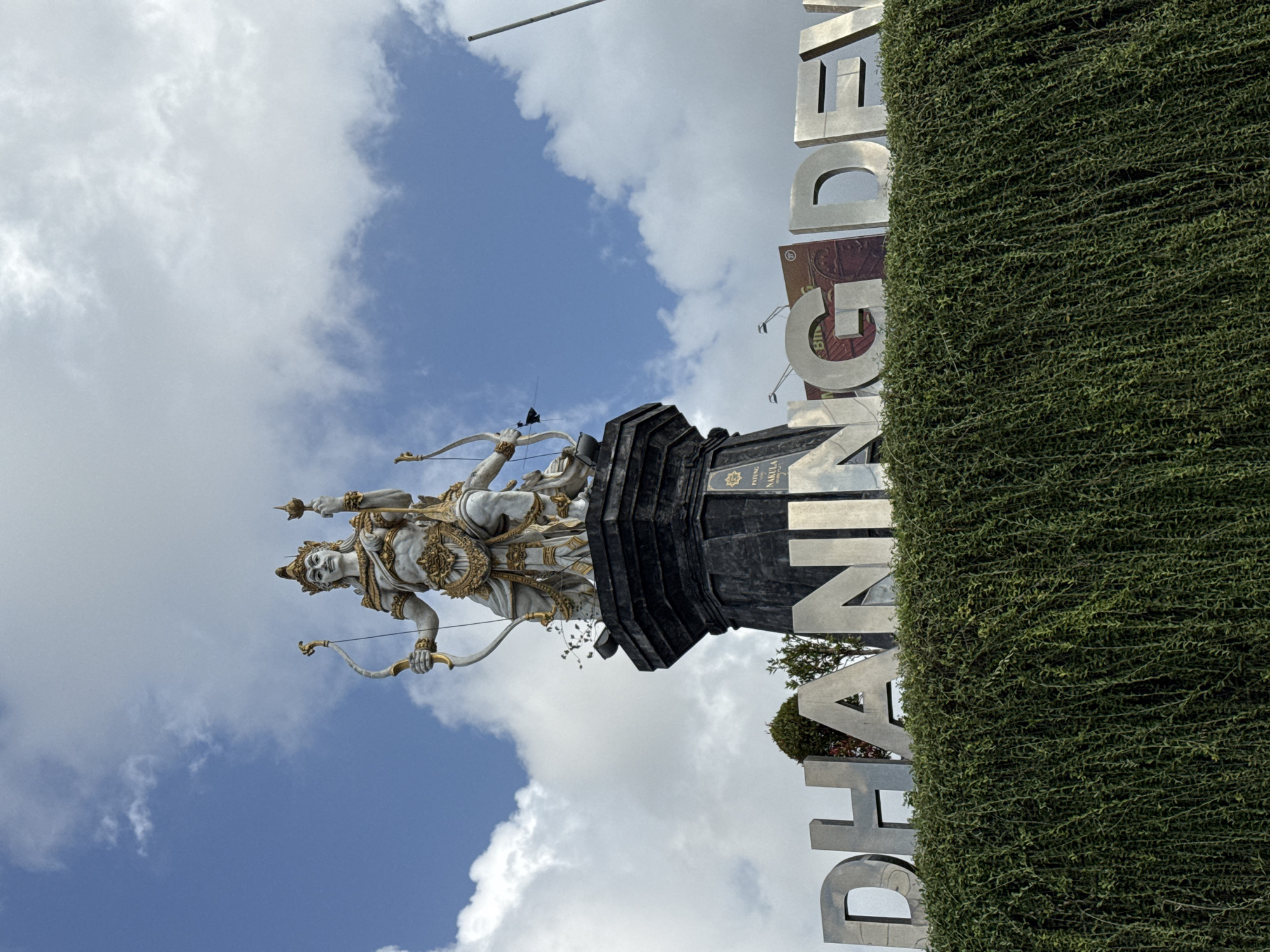 A statue of a Hindu deity holding a bow and arrow stands tall against a blue sky with scattered white clouds. The deity is adorned with golden ornaments and a crown. Below the statue, large silver letters spell out "DHANING DEV". The letters are partially obscured by a dense wall of green, hanging foliage. In the background, billboards and a pole are visible. The scene is located in Kuta Selatan, Indonesia, as indicated by the text.