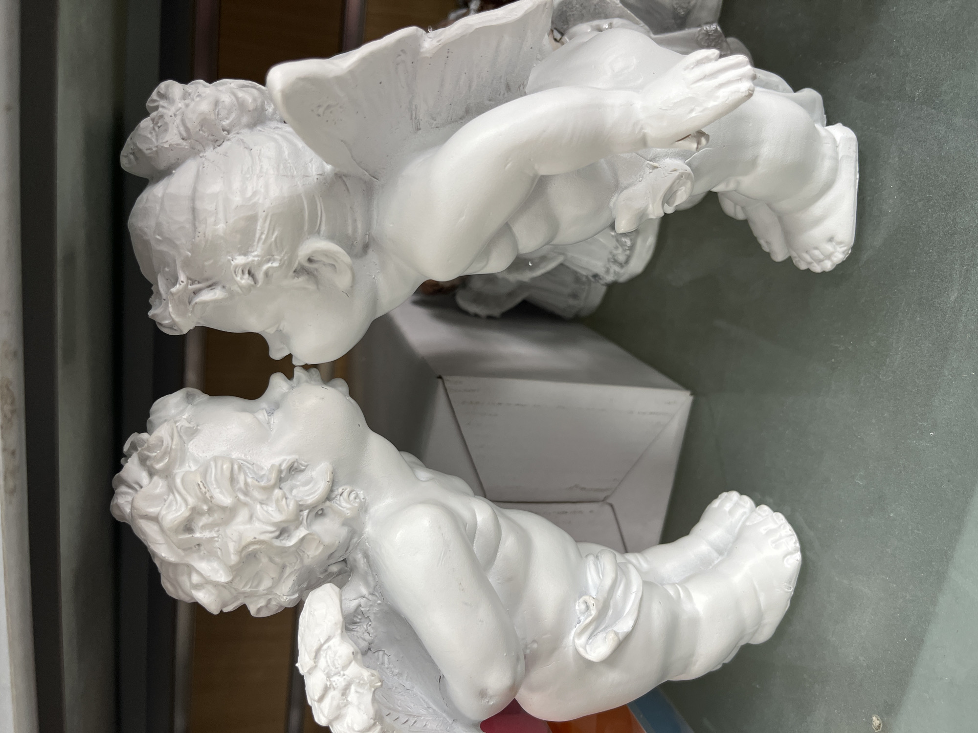 Two white, plaster cherubs are positioned near each other, appearing to face each other. A plain white box is situated between them, partially obscured. The background appears to be a dull, neutral surface. The specific location in the city of Budva, Montenegro, is not discernible from this image.