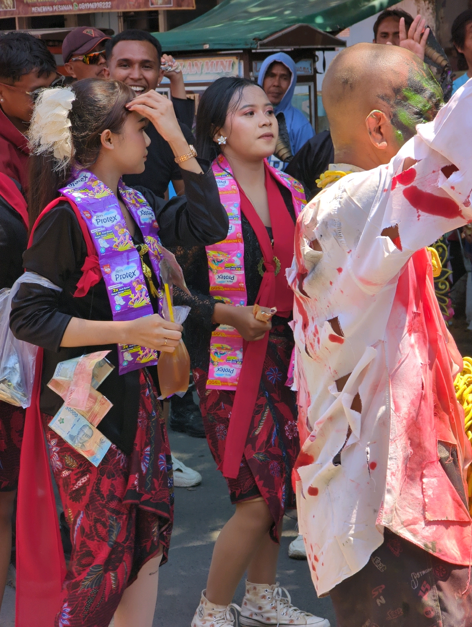 In a street scene in Pasuruan, Indonesia, likely during a festival or event, three young adults are the central focus. On the left, a woman with a white hair accessory and a black outfit wears a sash made of Protex soap sachets. She holds a plastic bag with money, including Indonesian rupiah denominations of 50,000 and 100,000, and a drink in a plastic pouch with a yellow straw. To her right, another young woman in a black top and a patterned red and black skirt is also adorned with a similar Protex sachet sash and a red scarf. She is looking to her right. On the far right, a person with a shaved head, painted green and black on their scalp, wears a tattered white garment stained with red, resembling blood, suggesting a costume or performance, possibly a zombie or horror theme. The background shows other people, including a man in a blue hoodie and a man with a hand raised, and the environment suggests an outdoor market or street event with awnings and stalls visible. The ground appears to be asphalt.