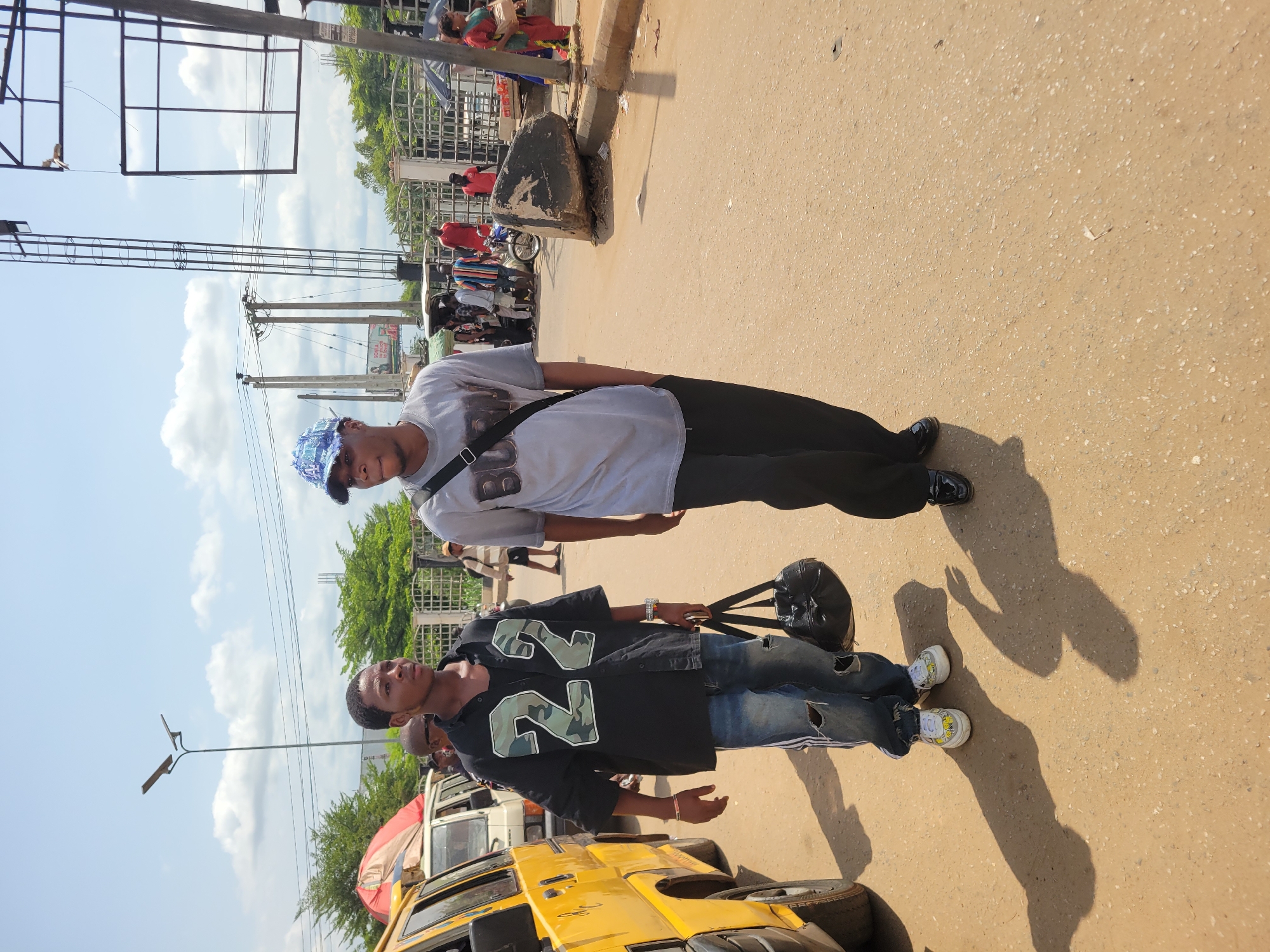 The image depicts a bright, sunny street scene, likely in an urban area consistent with Onitsha, Nigeria. Two young men stand in the foreground on a light brown, somewhat granular road surface.
The man on the left wears a black short-sleeved shirt featuring a large "22" in a camouflage pattern, ripped blue jeans, and white sneakers with yellow details. He carries a black shoulder bag and has a watch on his left wrist. His gaze is directed slightly to his right.
The man on the right is dressed in a light grey t-shirt with a dark graphic design that includes a prominent "B" or "8", black trousers, and shiny black dress shoes. He wears a blue patterned baseball cap and has a black shoulder bag slung across his body. He looks towards his left.
Both individuals cast long, distinct shadows on the ground, indicating a high sun angle, suggesting it is midday or early afternoon. The sky above is clear with some scattered white clouds.
In the background, several other people are visible, some walking and others standing near vehicles. A yellow commercial bus or taxi is partially seen on the far left. Utility poles with multiple overhead wires crisscross the sky, alongside a large, empty metal billboard frame. Green trees and metal fencing or gates line the background. A concrete curb and a large rock are also visible along the side of the road. The scene portrays a typical bustling street environment with general pedestrian and vehicular activity.
