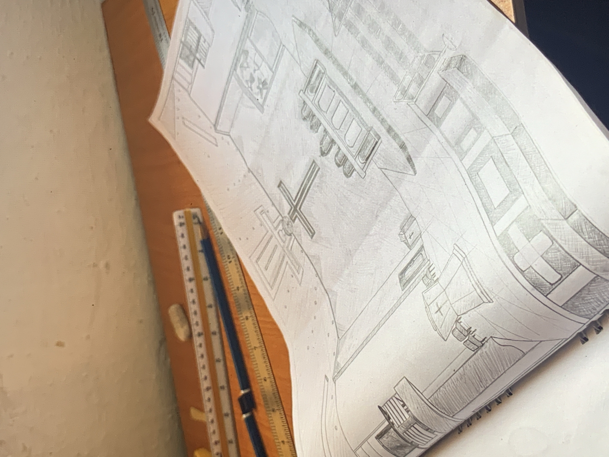 The image displays a detailed pencil drawing on white paper, laid out on a light brown drafting board or table. The drawing appears to be a technical or architectural plan, likely depicting the interior layout of a building, possibly a church or a large hall.
In the center of the drawing, a prominent cross-shaped structure is visible, surrounded by other geometric forms. To the upper left of the cross, there is a long, rectangular object with several smaller, segmented rectangles within it, resembling rows of pews or tables with chairs. Additional rectangular and linear elements throughout the drawing suggest walls, windows, and other architectural features, some shaded with cross-hatching to indicate depth or texture. The paper is slightly curled and shows signs of use, typical of a work in progress.
To the left of the drawing, a set of drafting tools is present on the brown surface. These include a black pencil with a metallic clip and an oval-shaped white eraser. Beneath the pencil, a long, clear ruler or parallel rule arm with numerical markings is visible, suggesting precise measurement and straight lines were used in the creation of the drawing. The paper itself is spiral-bound on the right side, indicating it is part of a sketchpad or design notebook.
The scene is lit from the left, casting soft shadows and illuminating the textured surface of the paper and the drafting board. The overall impression is that of a workspace where a detailed design or illustration is being created or has recently been completed. No discernible text beyond the faint numerical markings on the ruler is clearly visible.