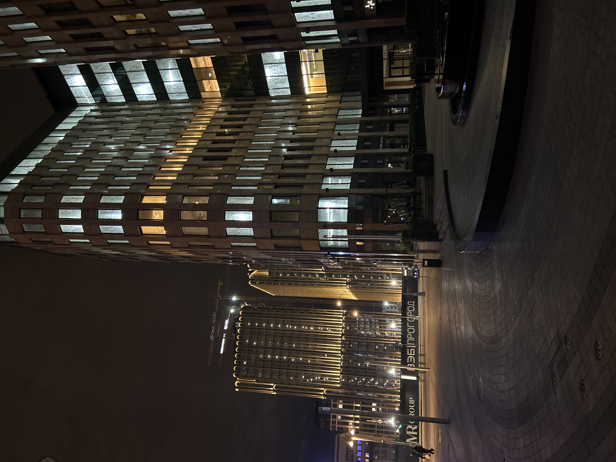 Captured at night, the image features two towering buildings, the tops of which are outside the frame. The taller of the two buildings is on the left, with illuminated windows and a grid-like facade. To its right is a shorter building, its facade defined by a series of vertical illuminated lines. A street or paved area occupies the foreground. Signs are visible, featuring text in a Cyrillic script, and some English text. The lighting suggests an urban setting with streetlights adding to the ambient luminescence.