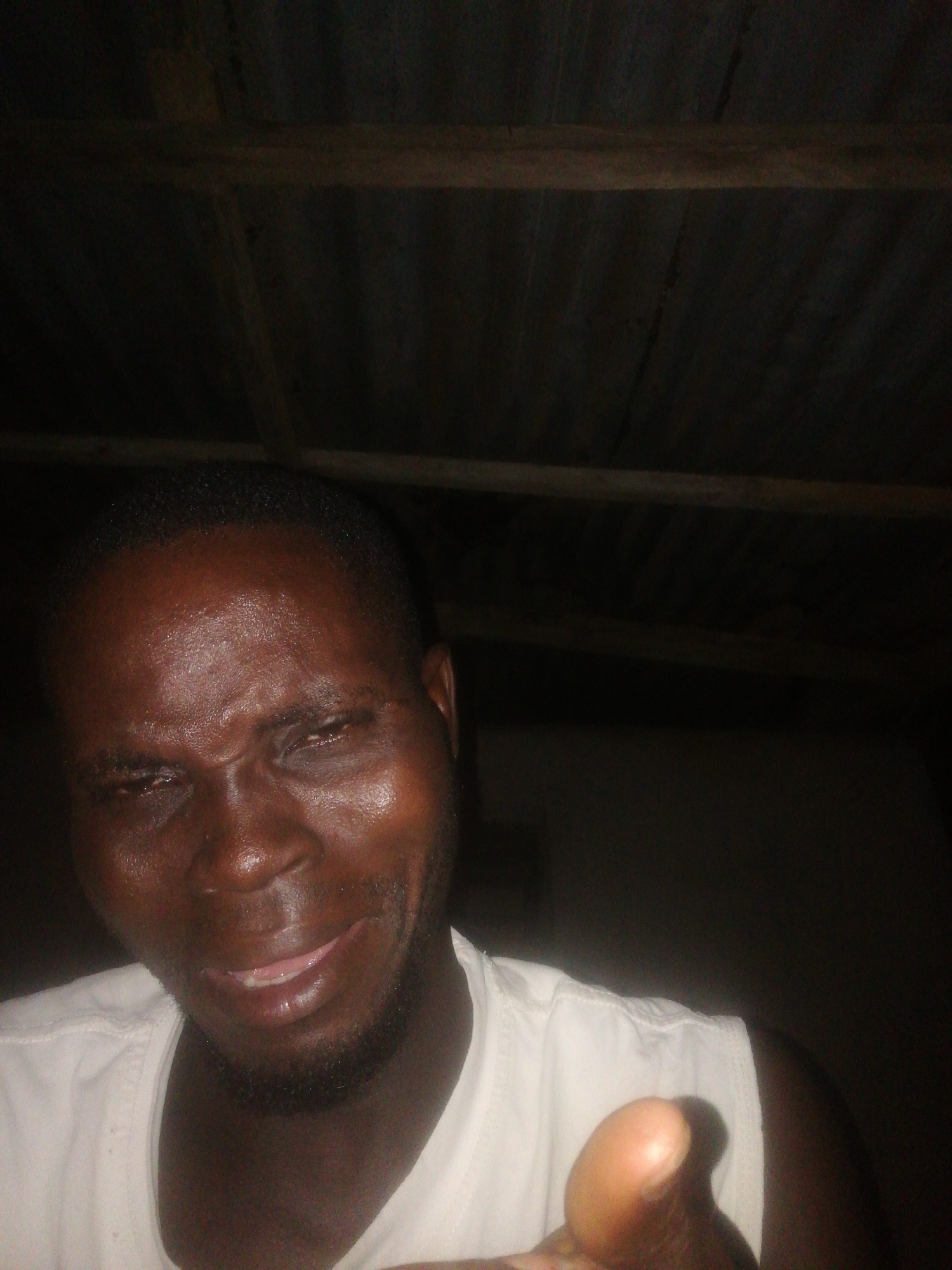 A man is in a dimly lit room, possibly indoors given the wooden ceiling visible at the top. The man has dark skin, close-cropped hair, and a short beard. He is wearing a white sleeveless shirt. He is pointing directly towards the camera with his right index finger extended. The environment is dark, making it difficult to discern details. There is no evidence in the image to confirm the location of Ikorodu, Nigeria.