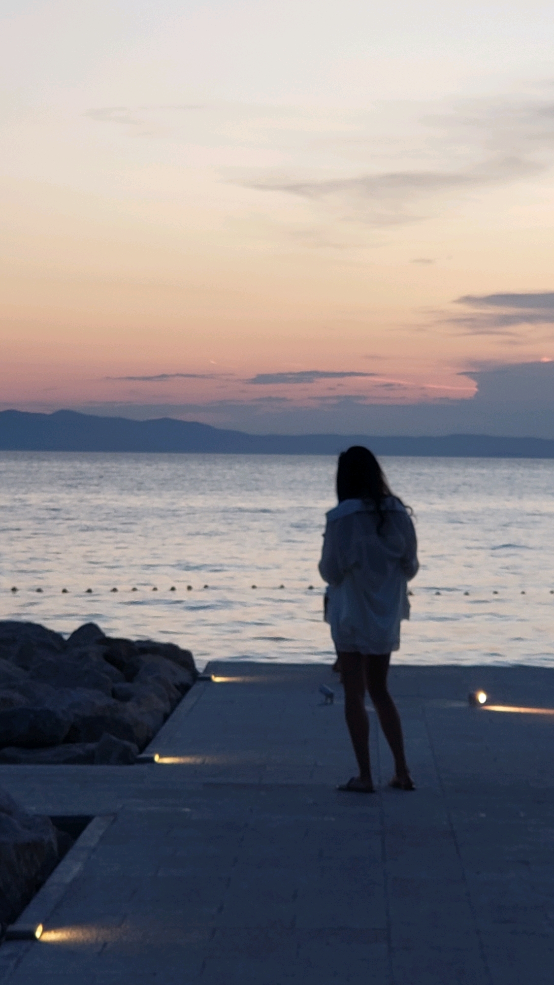 A person stands on a paved walkway overlooking a body of water, likely the Adriatic Sea, with a silhouetted mountain range in the distance. The sky displays a gradient of pastel colors, indicating either sunrise or sunset. The walkway is illuminated by several small, recessed lights. To the left of the walkway, large rocks form a breakwater. The person, facing away from the camera, is wearing a light-colored jacket and shorts.