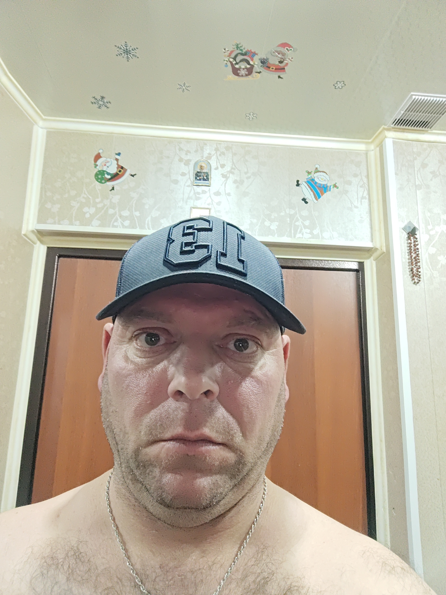 A man with fair skin, stubble, and a light complexion is pictured in a frontal, close-up shot. He is wearing a dark baseball cap with a prominent "EJ" logo. His expression is neutral, with his mouth closed and eyes open. The background consists of a door and wallpaper adorned with small, festive holiday-themed stickers of Santa Claus and snowflakes. The ceiling also features similar decorative stickers. A silver chain is visible around the man's neck. The overall impression is a casual, indoor setting, possibly a bathroom or bedroom, with a hint of holiday decoration. The location is identified as Polyarny, Russia.