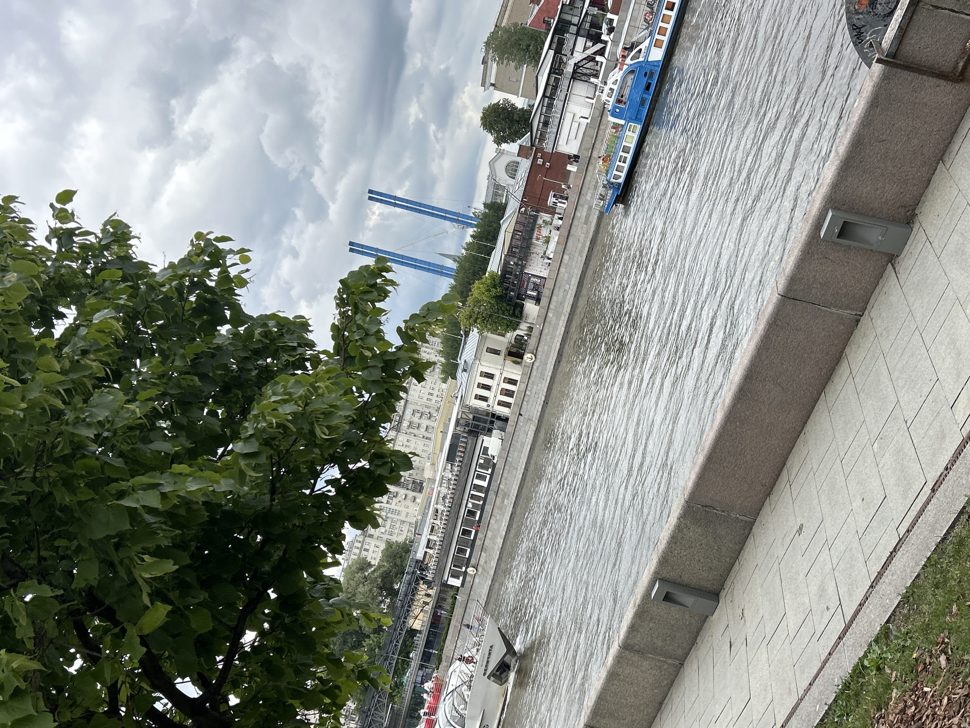 The image shows a river scene in an urban environment. The river occupies a significant portion of the frame, its surface reflecting the cloudy sky above. A tree with dense green foliage is prominent on the left side.
Along the riverbank, a paved walkway runs adjacent to the water's edge. Buildings of varying architectural styles line the riverbank. There are passenger boats docked along the quay. In the distance, blue supports for a bridge-like structure are visible.
The overall scene suggests a waterfront area in a city.