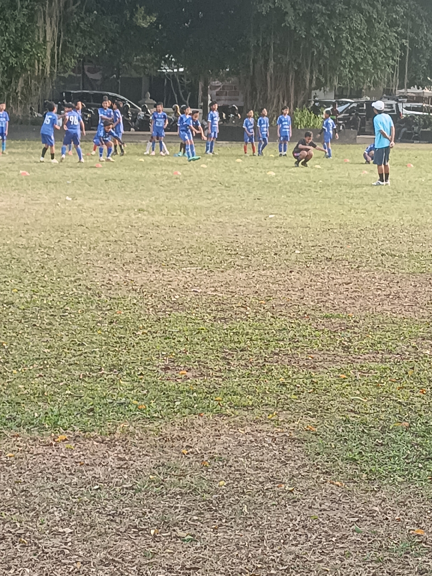 This is a medium shot, outdoor, eye-level, full color, slightly blurry image taken on a cloudy day in Sleman, Indonesia. The scene depicts a group of children, likely a youth soccer team, on a grassy field. Several children, dressed in blue soccer uniforms with white accents and some with numbers visible on their jerseys, are scattered across the field. Some are in motion, appearing to be participating in a drill or practice. One child in the foreground is squatting down, seemingly interacting with a coach or another player. Another adult, also in a blue shirt and shorts, stands facing away from the camera on the right side of the frame. In the background, trees with hanging roots and some parked vehicles are visible, suggesting the field is located in an urban or semi-urban park area. The ground is covered in patchy grass with some exposed soil and fallen leaves. The overall impression is one of a youth sports activity taking place in an open, outdoor setting.