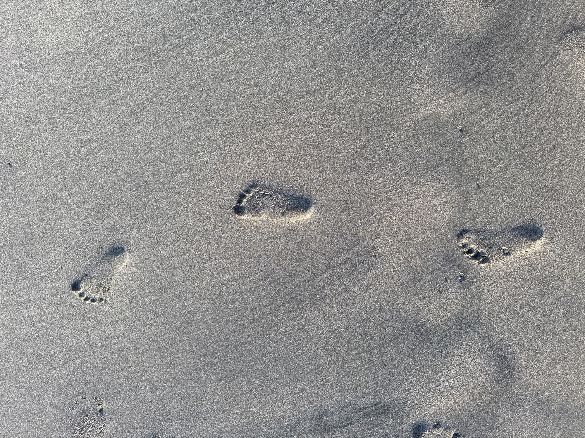 Footprints in the sand in Kuta, Indonesia. Three distinct footprints are visible, each showing the impression of toes. The sand is light gray and appears slightly damp. Faint striations run across the sand's surface. No people or other objects are visible in the scene.