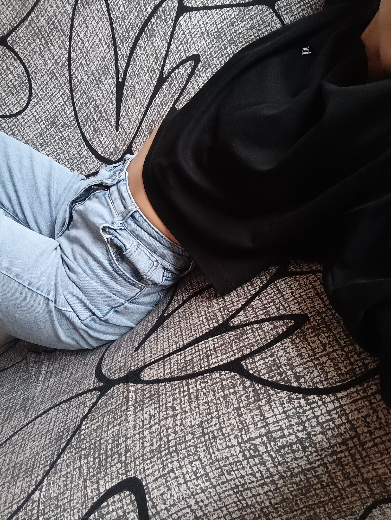 The image shows a person reclining on a patterned surface, likely a couch or bed. The person is wearing light-wash denim jeans and a black long-sleeved shirt. A portion of their torso is visible between the jeans and shirt. The patterned surface is a mix of grey, white, and black with a leaf-like design. The person is lying down and their body appears relaxed. There are no explicit indications of the location of the city of Chaikovsky, Russia. The image composition includes a tight focus on the body, excluding the face and any clear identifiers of setting, therefore precise location cannot be ascertained using visual information alone.