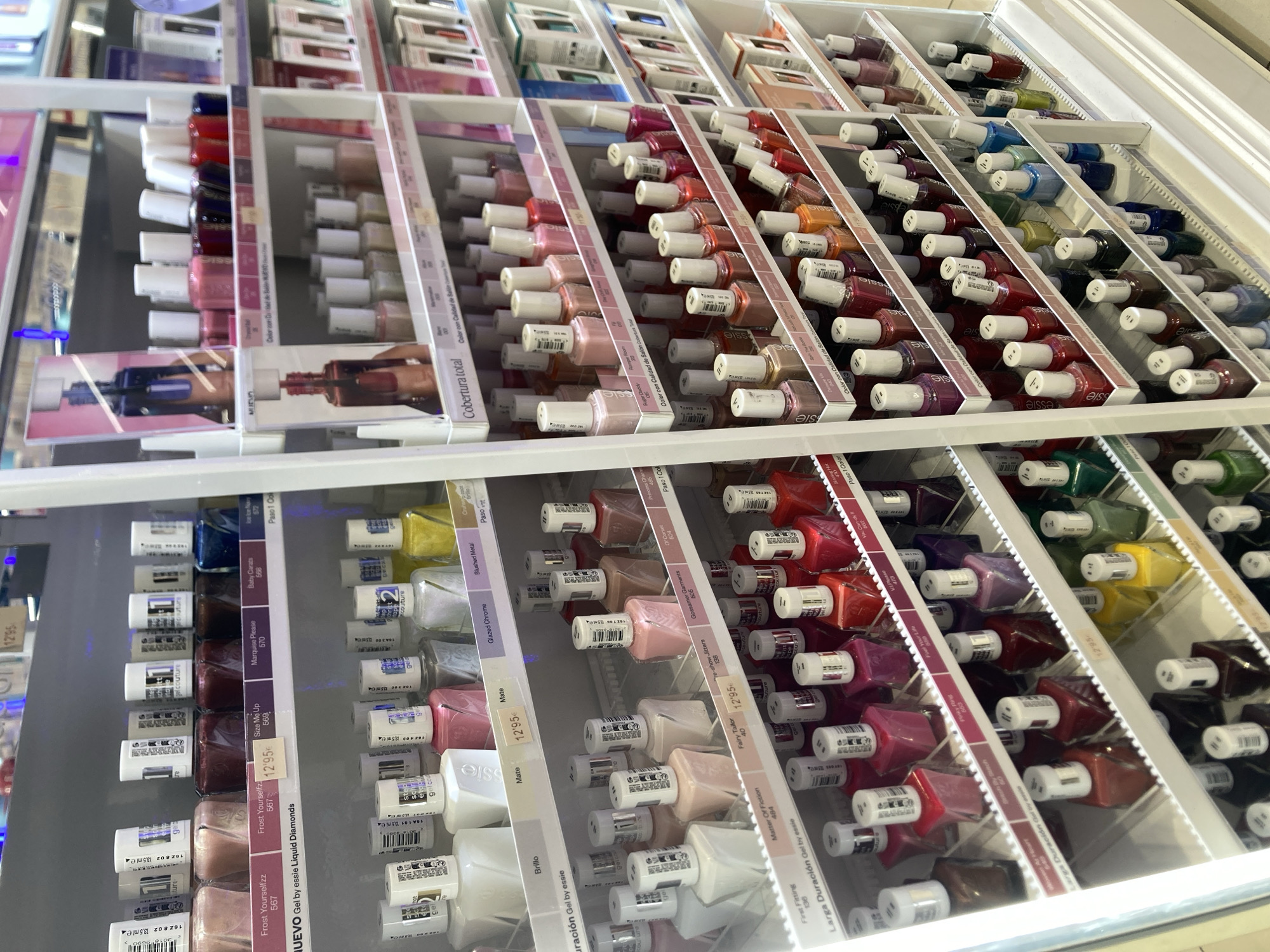 The image shows a retail display of Essie brand nail polish bottles. The display is located in a store in Barcelona, Spain, and features multiple shelves filled with rows of nail polish bottles of various colors. Some bottles are upright, while others are tilted or angled. Labels with product names and prices are visible on some of the shelves. The lighting is bright, suggesting an indoor retail environment. There are no people visible in the image.