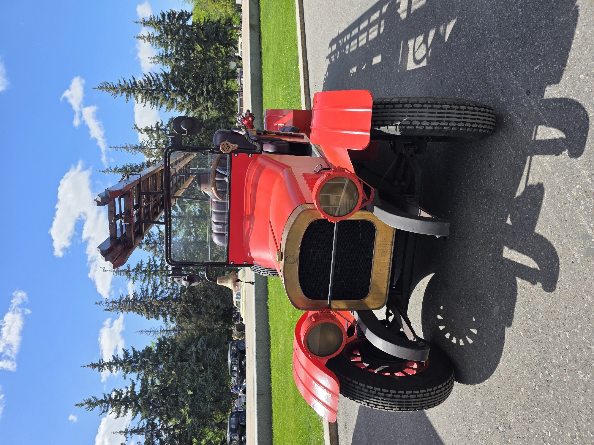 The image shows a vintage red fire truck parked on a paved surface. The truck has a wooden ladder mounted on the back. The surrounding environment includes green grass, trees, and a blue sky with white clouds. Based on the presence of vintage cars and the style of the fire truck, the location is likely a vintage car show or a historical reenactment event. While the city is requested, no definitive details directly link the location to Ufa, Russia.
