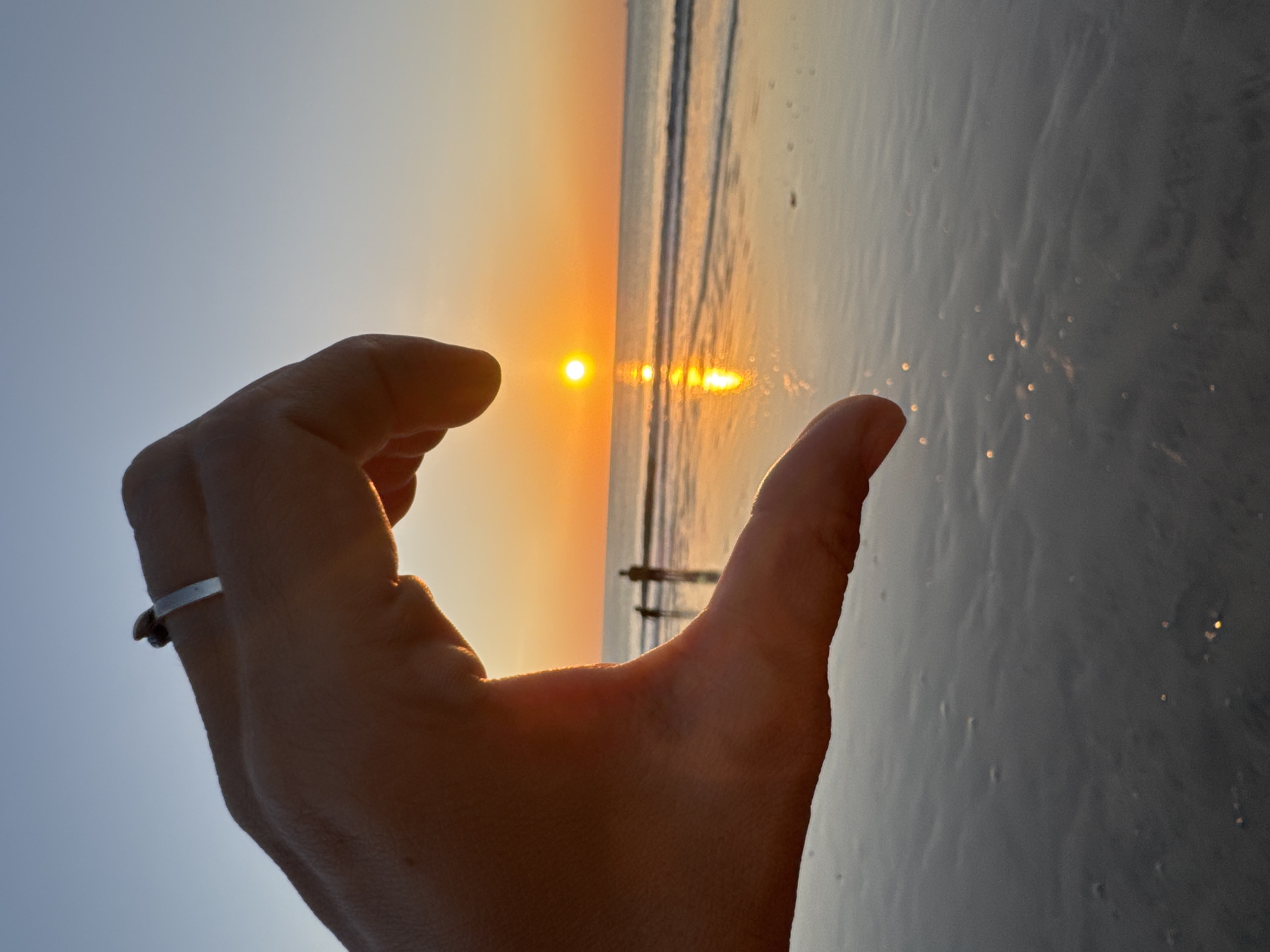 An extreme close-up shot focuses on a human hand forming a heart shape against a sunset backdrop over the ocean. The hand, positioned in the foreground, is silhouetted by the warm glow of the setting sun. A ring is visible on the index finger of the hand. In the background, the sun is setting over a calm ocean, casting a golden reflection on the water's surface. Two indistinct figures, likely people, are visible standing in the shallow water near the shore. The sky transitions from a clear blue at the top to a vibrant orange and yellow near the horizon, indicative of a sunset. The image appears to have been taken at a beach location, possibly in Kuta Selatan, Indonesia, known for its scenic sunsets and coastal views. The overall scene evokes a sense of romance and tranquility.