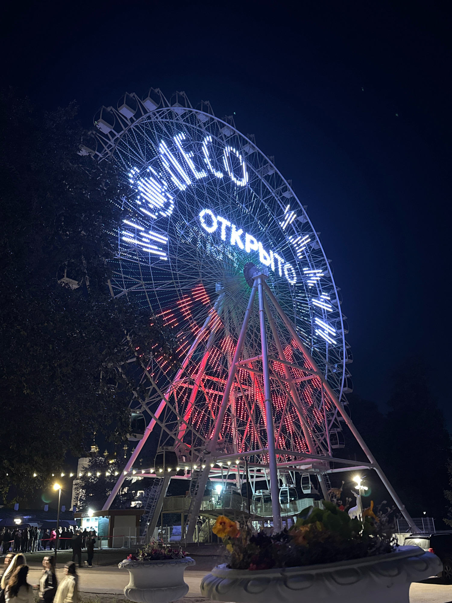 The image shows a Ferris wheel at night, illuminated with white and red lights. The Ferris wheel is located in Tver, Russia, specifically in the city park. The white lights on the Ferris wheel spell out "WELCOME" and "ОТКРЫТО," which translates to "OPEN."
In the foreground, there are people standing and walking around, and there are also planters with flowers. In the background, there are more lights from buildings and trees. The sky is dark, indicating that it is nighttime. The overall scene depicts a lively atmosphere in a park setting at night.