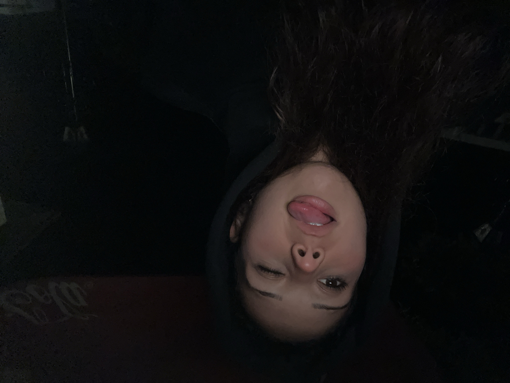 A young woman with dark, wavy hair and a black hooded sweatshirt winks at the camera, sticking her tongue out slightly. She appears to be in a car at night. The background is dark, but a faint outline of a Coca-Cola logo is visible on the right side of the image. The lighting suggests the photo was taken with a flash. There is no information in the image that suggests a location in Zlatograd, Bulgaria.