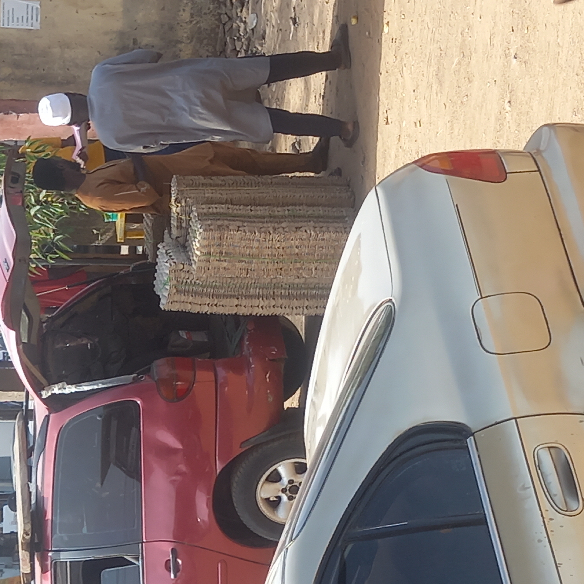 The image captures a bustling street scene in Sardauna, Nigeria, under bright daylight. The central focus is on a commercial activity involving a large, tall stack of woven mats, likely made of dried grass or reeds, bound together with green strands.
Two men are visible, appearing to be engaged in the activity. One man, seen from behind, wears a white skullcap and a light-colored long tunic over dark trousers. He stands near the large stack of mats. Another man, wearing an orange or light brown tunic, stands partially obscured behind the stack of mats. A third individual, largely out of frame, is visible to the left of the man in the orange tunic, wearing a yellow and green patterned head covering.
The stack of mats is positioned close to the rear of a red hatchback car, which has its trunk open, suggesting the mats are being loaded, unloaded, or displayed for sale. Items are visible inside the open trunk of the red car. In the immediate foreground, a light-colored sedan (possibly silver or champagne) is partially visible, showing its rear quarter panel, taillight, a fuel cap outline, and a door handle.
The ground is unpaved, consisting of packed dirt and small stones. In the background, there is a light-colored building wall and some green foliage. The bright sunlight indicates clear weather. A small, white rectangular paper with faint, illegible text is visible on the background wall. The scene depicts a typical commercial exchange in an outdoor, local setting.