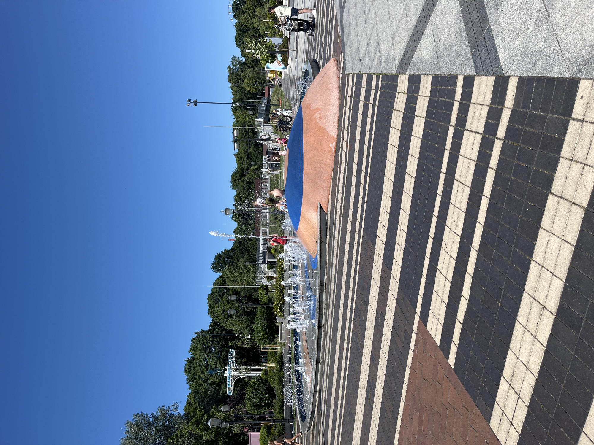 The image depicts a sun-drenched public space in Kaliningrad, Russia, likely a park or promenade, on a clear day. The scene is dominated by a modern fountain with water jets spraying upwards, suggesting it's a recreational area for warm weather. The ground is paved with contrasting tiles, creating a striking striped pattern that casts long shadows, indicating it is likely late morning or early afternoon.
Several people are visible, engaged in leisure activities. In the foreground, a person in swimwear is near the fountain. Further back, a child with a white helmet is playing in the water, and other individuals can be seen around the fountain area. A person on a bicycle is also present, adding to the sense of a public gathering space.
The background features lush green trees and some structures, including lampposts and what appears to be a viewing tower or attraction. The clear blue sky and bright sunlight suggest pleasant weather, inviting outdoor activities. The overall mood is one of enjoyment and relaxation in a well-maintained urban environment. No visible text is present in the image.