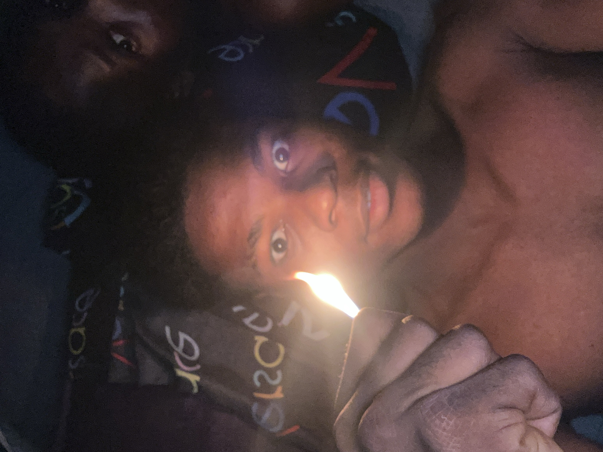 The image shows a close-up of a young Black male holding a lit lighter in the foreground, with his face illuminated by the flame. The background appears to be a dark surface with colorful lettering or patterns. The man's eyes are wide open, and he is looking directly at the camera. His upper body is bare. The overall lighting is dim, with the flame of the lighter providing the primary light source. No specific location in Ihiagwa, Nigeria, is depicted or identifiable.