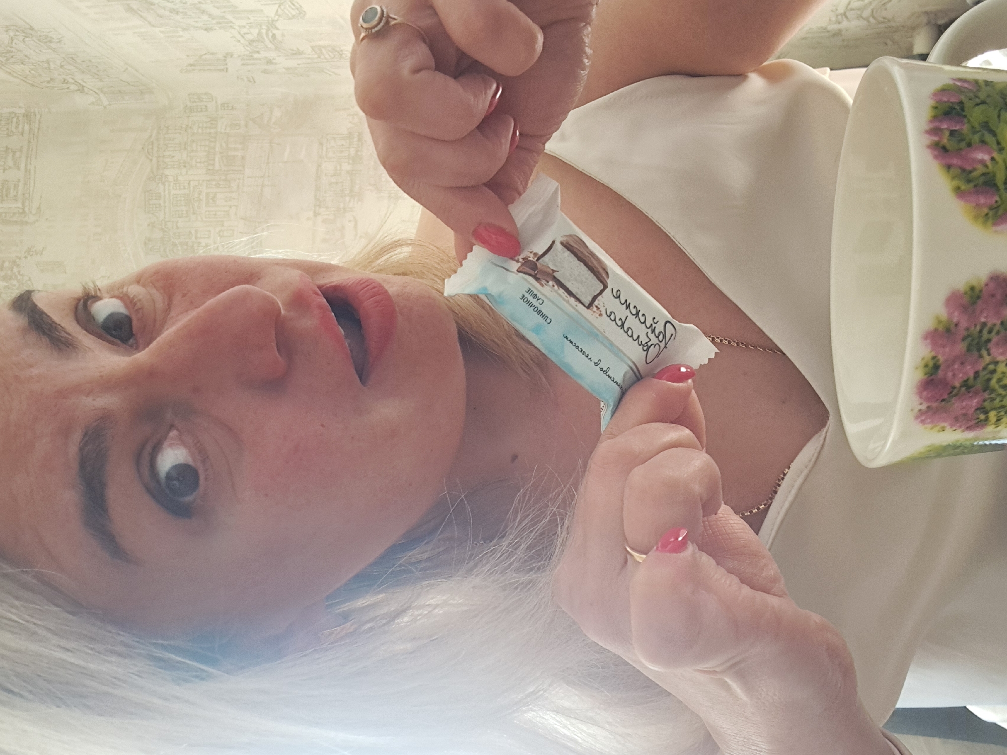 A Caucasian woman with fair skin and graying blonde hair is positioned in the foreground, holding a packaged dessert and a floral-patterned mug. She is wearing a white top and a thin gold necklace. The background consists of a light-colored wallpaper with a pattern of city blocks. The package in her hand is labeled with Russian text. Given that the location is Elektrostal, Russia, the writing on the packaging is most likely in Russian.