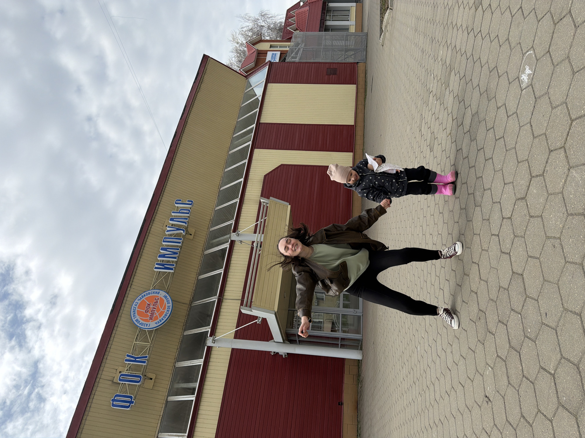 A woman and a child stand outside the building of the "FOK IMPULS" sports complex in Chaikovsky, Russia. The building is two stories tall with a facade of alternating horizontal bands of beige and red paneling. The name of the sports complex, "ФОК ИМПУЛЬС", is displayed in blue lettering above the entrance. The woman wears a brown jacket, green shirt, and black pants with white sneakers. She has dark hair and is smiling. The child wears a dark jacket with a dotted pattern, black pants, pink boots, and a light pink hat. The sky is overcast. The ground is paved with hexagonal concrete blocks.