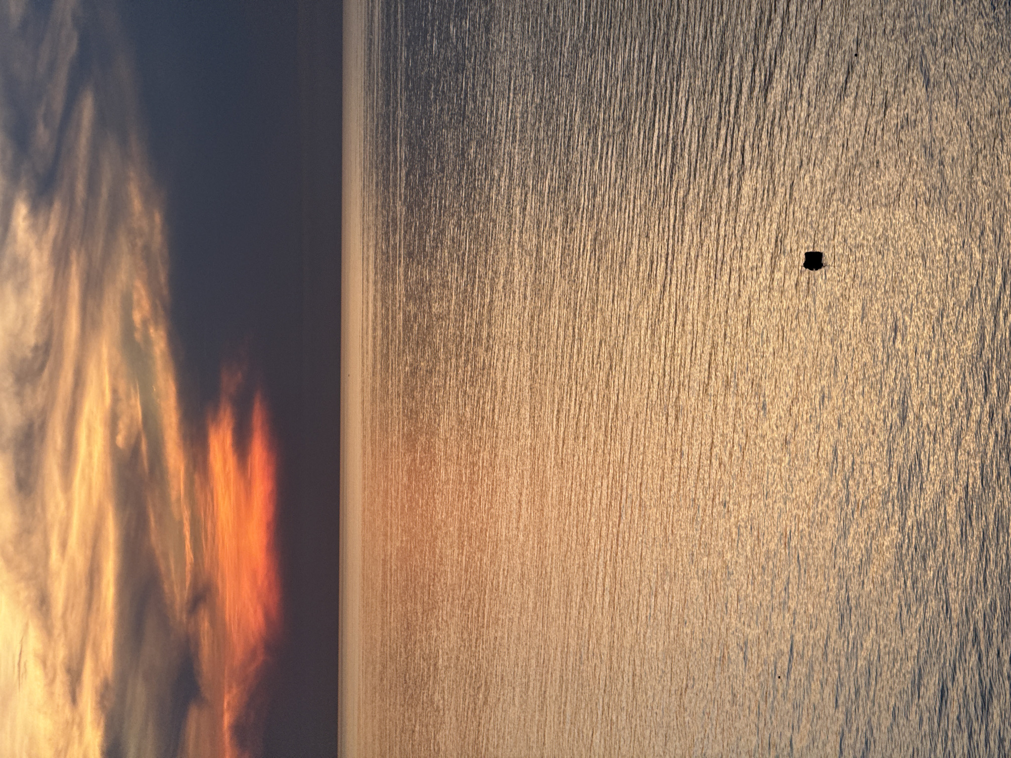 The image depicts a serene seascape at sunset, as seen from Lý Sơn Special Administrative Region, Vietnam. The sky is painted with a vibrant palette of colors, ranging from fiery orange and yellow near the horizon to deeper shades of purple and dark gray towards the top. Wispy clouds are scattered across the sky, catching the last rays of the sun. The sea is calm, reflecting the warm hues of the sunset, with gentle ripples creating a textured surface. A small, dark object, possibly a buoy or a piece of debris, floats in the middle of the water. The overall atmosphere is tranquil and visually striking.