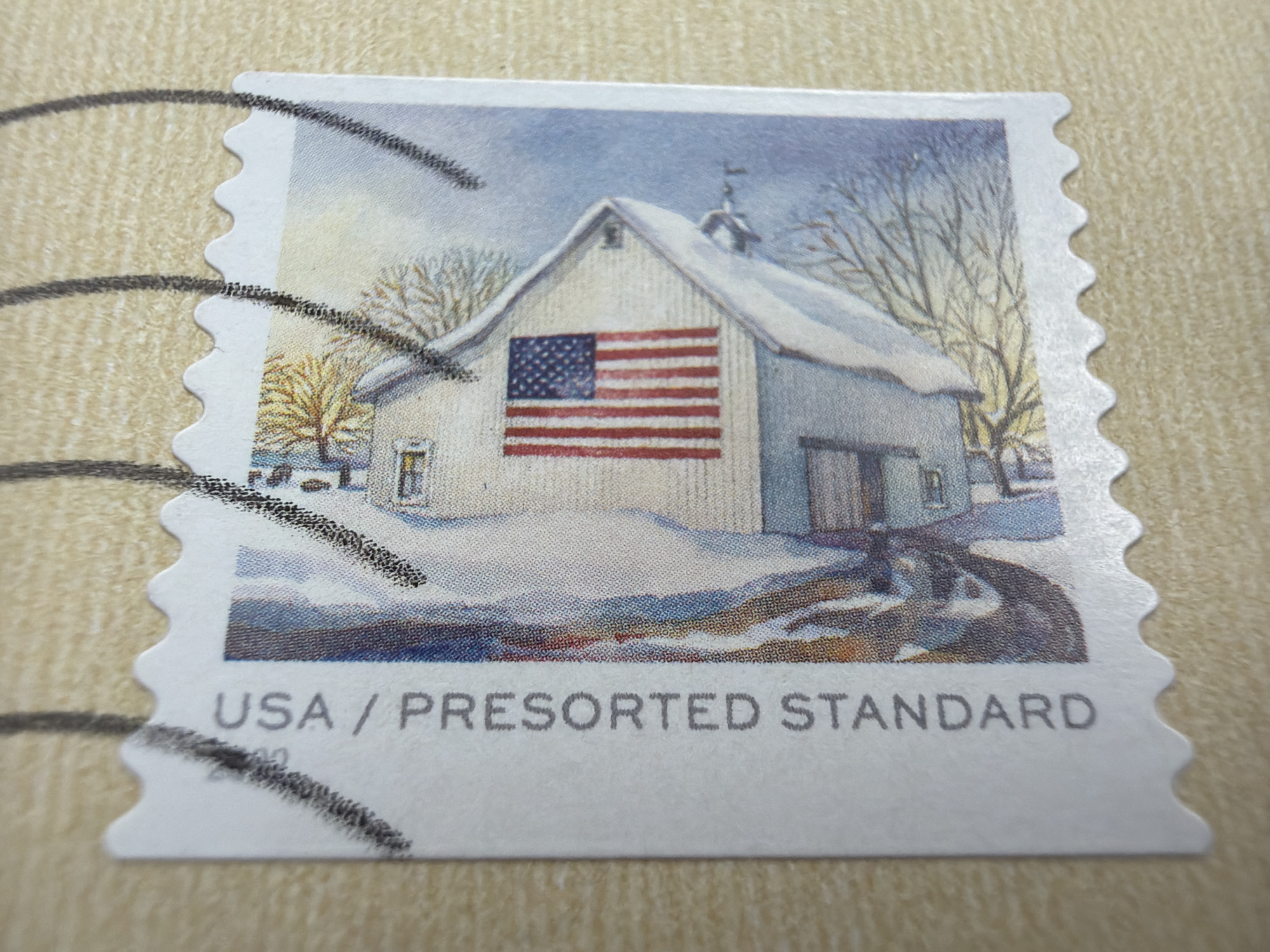 The image shows a close-up of a postage stamp. The stamp features a watercolor illustration of a white barn covered in snow, with an American flag painted on its side. Bare trees and a winter landscape surround the barn. The bottom of the stamp reads "USA / PRESORTED STANDARD" in gray text. The stamp is placed on a textured, light brown surface. There is no indication of the city of Grapevine, United States of America, in the image.