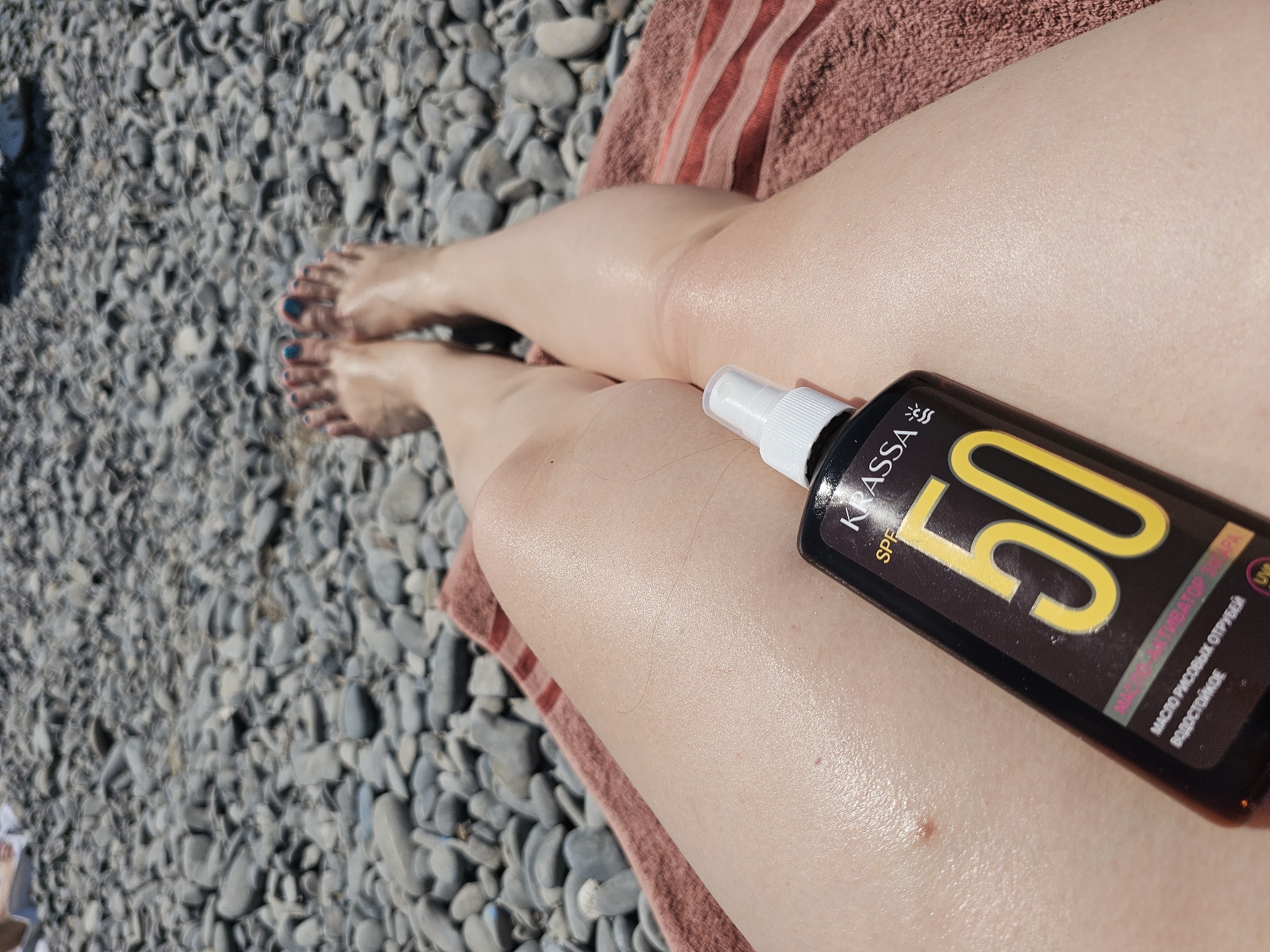 An aerial perspective shot shows a person's legs and feet resting on a towel on a pebble beach. The person's toes are painted with blue nail polish. A spray bottle of "KRASSA SPF 50" sunscreen is placed on the person's thigh. The beach appears to be in Kirillovka, Russia. The background is filled with grey and white pebbles. The lighting suggests it is daytime, likely sunny.