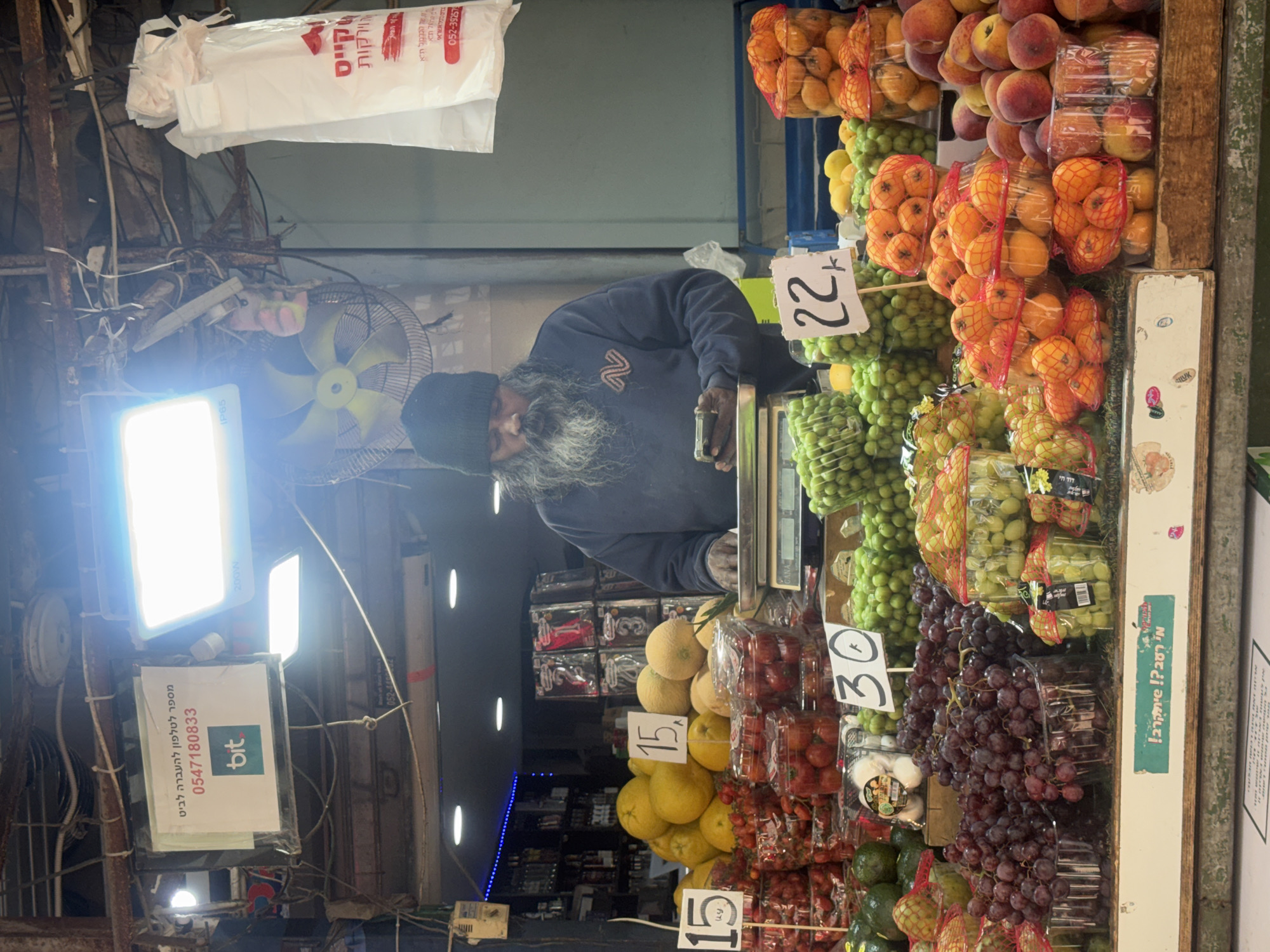 An image taken at a market in Tel Aviv, Israel, depicts a man with a long grey beard wearing a black beanie, standing behind a fruit and vegetable stall. He is weighing items on a scale. The stall is packed with produce, including grapes, oranges, peaches, melons, and other fruits, all marked with price signs in Hebrew. In the background, there is a mix of electrical wires, lights, and other market fixtures. A sign with Hebrew text and "bit" logo is visible. The lighting appears to be a mix of natural and artificial, with bright spotlights illuminating the area.