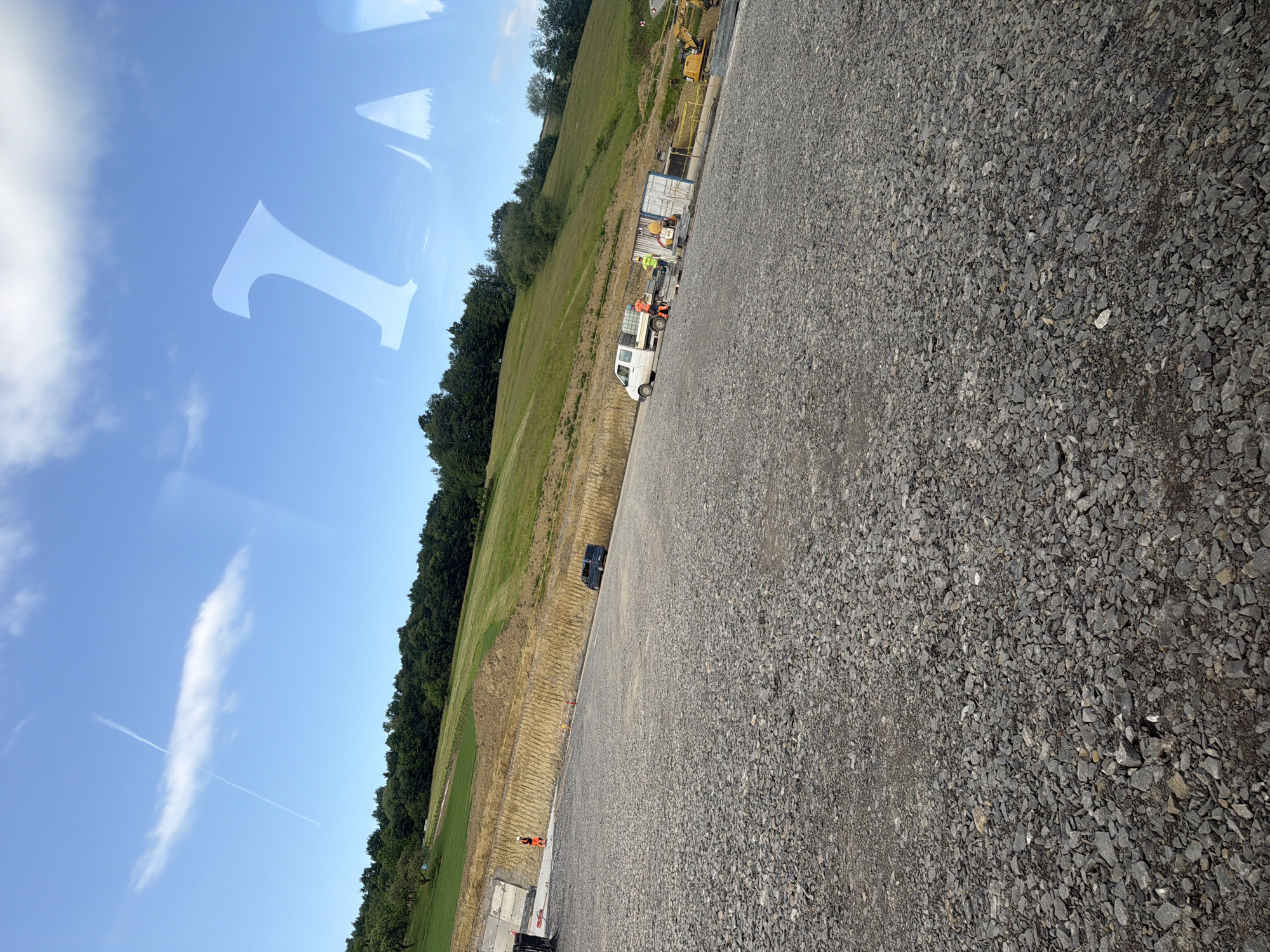 The image depicts a construction site in Stróża, Poland, characterized by a vast expanse of grey gravel in the foreground. In the midground, a sloped grassy hill rises, transitioning into a dense forest at the top. A construction worker, dressed in an orange high-visibility vest and white hard hat, stands near a white utility truck. Beside the truck, a white shipping container sits, with two construction workers in yellow high-visibility vests nearby, one appearing to be working with equipment. A yellow excavator is also visible further down the site. In the background, a clear blue sky is dotted with wispy white clouds, and a faint contrail from an aircraft is visible. The overall impression is one of ongoing construction work in a rural or semi-rural setting.