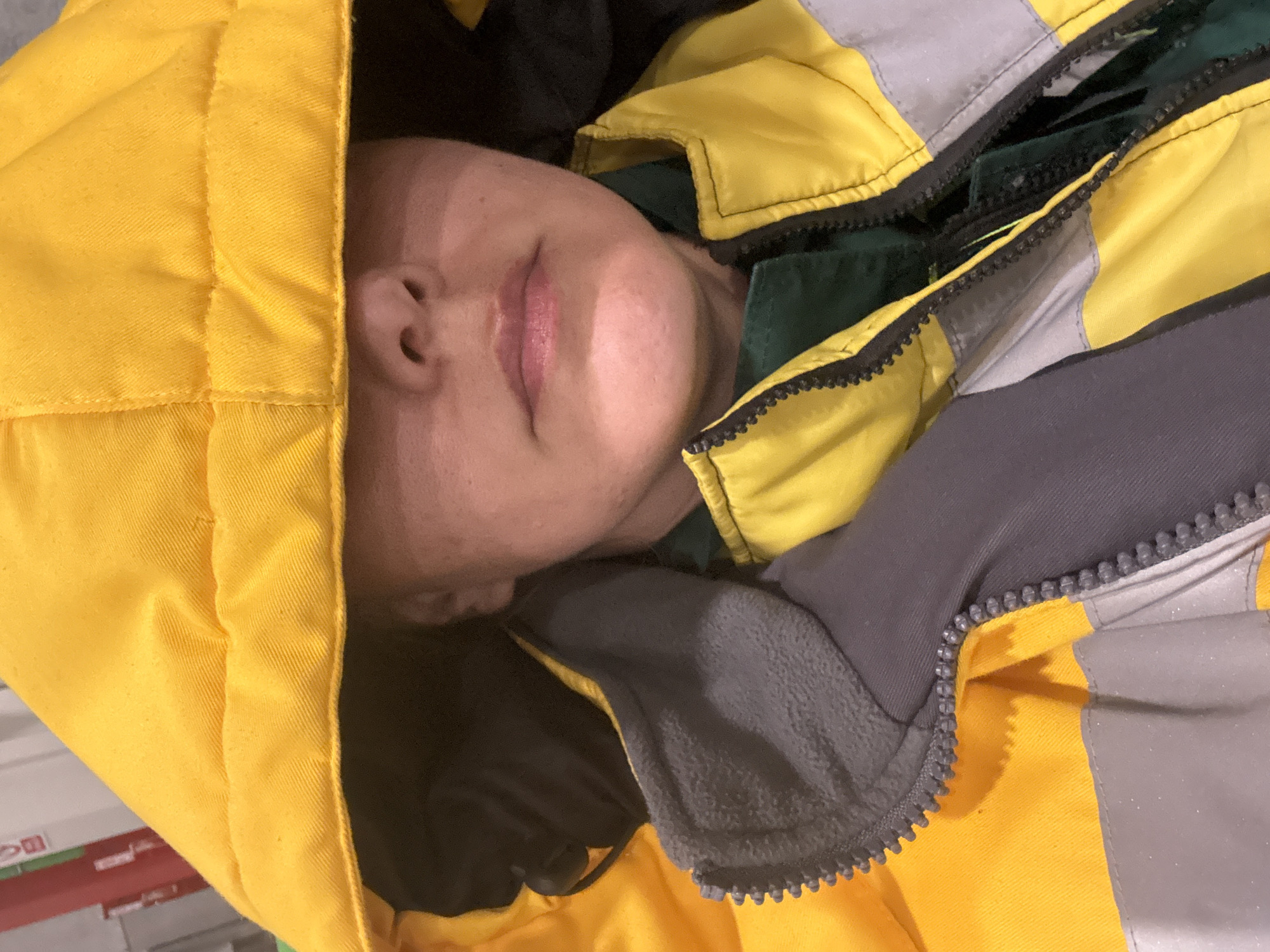 A person's face is visible, framed by a yellow hooded jacket. The jacket appears to be multi-layered with additional grey and black components. The person's eyes are closed, and their face is angled to the side. The location is unknown and there is no information available to suggest it is Lupanovo, Russia.