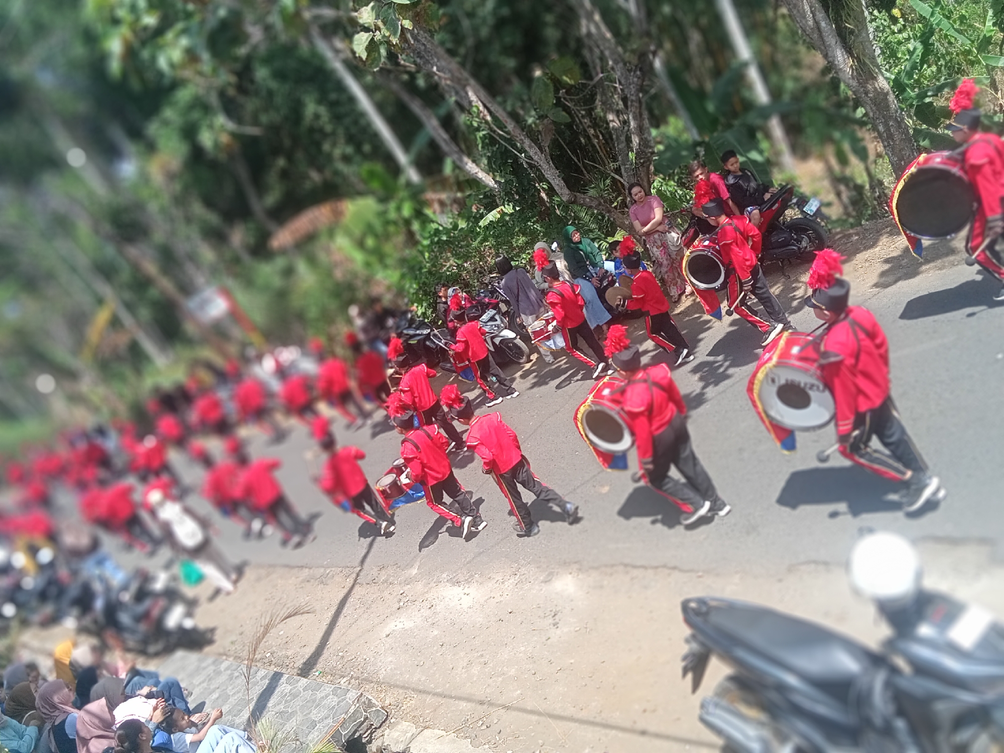 In Pule, Indonesia, a marching band in red and black uniforms with red feathered hats parades down a paved road bordered by lush greenery and trees. Several members are playing large bass drums, some with yellow and blue trim, while others carry smaller drums or are positioned to play them. Spectators, some seated on the side of the road, watch the procession. Motorcycles are parked along the road and on the embankment. The scene is illuminated by bright daylight.