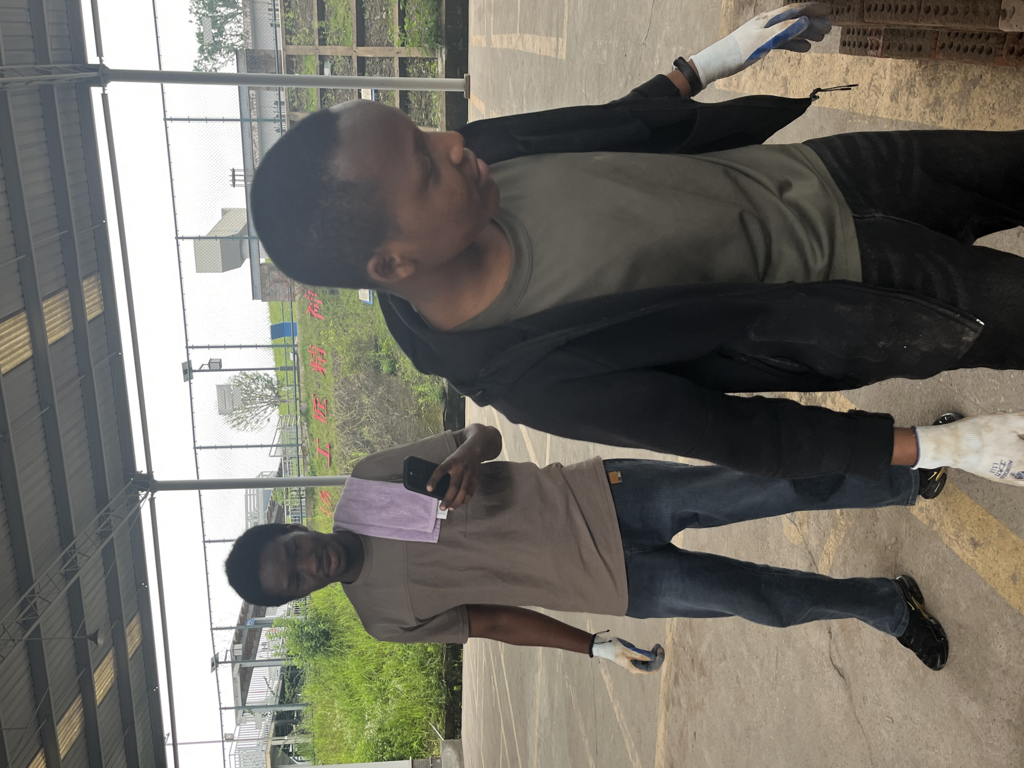 Two men are standing in an outdoor paved area with a covered roof overhead. The man on the left is wearing a brown t-shirt, blue jeans, black athletic shoes, and white and blue work gloves. He is holding a smartphone in his right hand and has a purple towel draped over his shoulder. The man on the right is wearing a green t-shirt, a black jacket, dark pants, and white work gloves. He is looking down and to his right. The background shows a chain-link fence, some greenery, and industrial-looking structures, including a large white ventilation duct. There are red Chinese characters visible on a grassy slope in the background. This appears to be an industrial area within the city of Jiangjin, China.