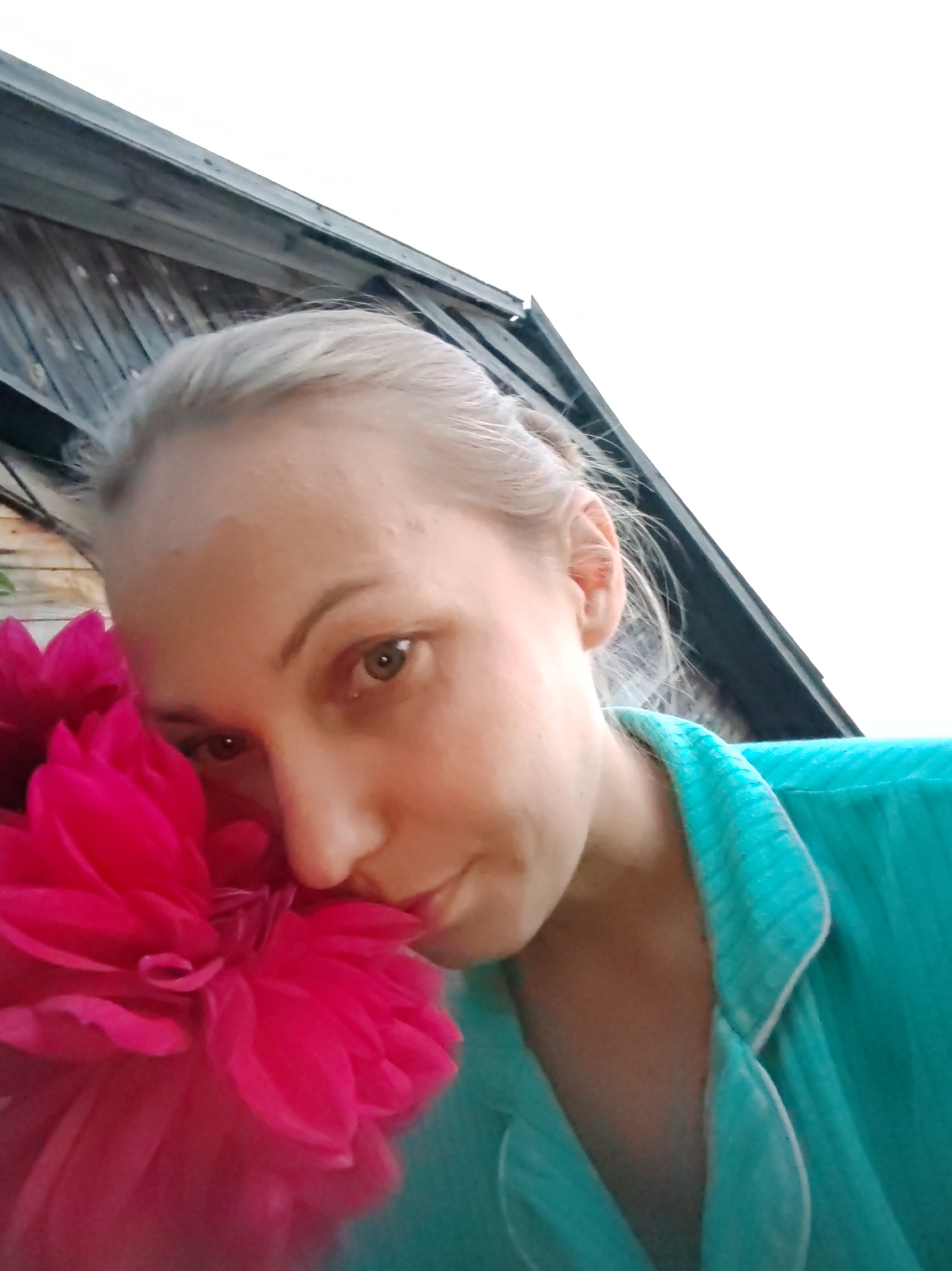 A close-up, low-angle shot shows a fair-skinned woman with blonde hair, looking slightly off-camera to her left. She is holding a large, bright pink dahlia flower close to her face, with the petals obscuring part of her left eye and cheek. The woman is wearing a teal-colored pajama top with a white collar. Behind her, a wooden structure with a dark, angled roof is visible against a bright, featureless sky. The overall scene is outdoors, possibly in a rural setting or a backyard, as suggested by the wooden building. The lighting suggests it is daytime, with the sky being overcast or overexposed. The image is sharp and well-lit, focusing on the woman and the flower.