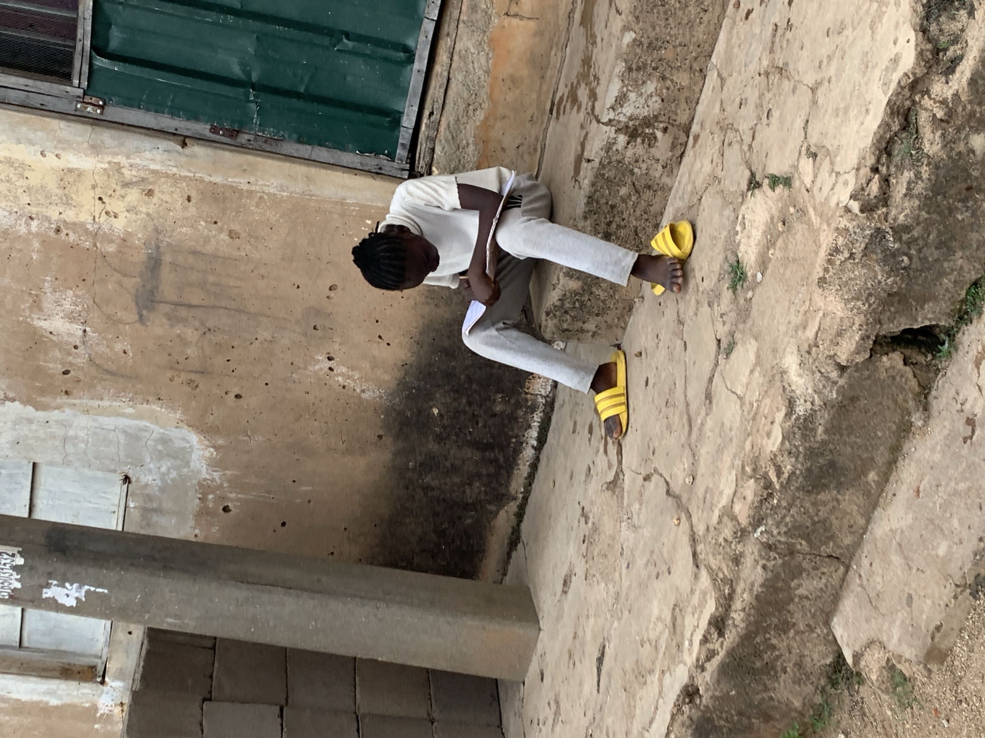 A young person is seated on a concrete platform beside a building in Jos, Nigeria. The person wears a light-colored long-sleeved shirt, gray pants, and yellow sandals. They appear to be studying or writing, holding a piece of paper in their hands. The building behind them has a green window or door. The concrete platform is cracked and shows signs of weathering. The environment seems to be urban, with a mix of concrete and vegetation.