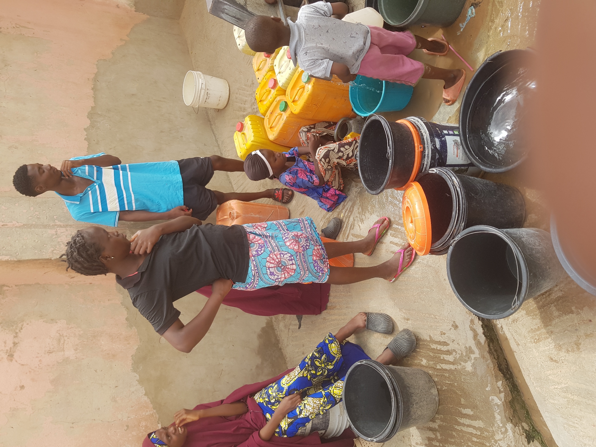 The image shows a group of individuals, including children and adults, positioned on a concrete or tiled surface in Dutsinma A, Nigeria. They are surrounded by yellow plastic jerrycans and several black buckets, some of which appear to contain water, indicated by the presence of reflections. One child is pouring water from a blue bucket. Some individuals are seated or lying down, while others are standing. The environment is slightly wet, suggesting a recent spillage. Clothes are colorful, and a white bucket is seen in the background near a wall.