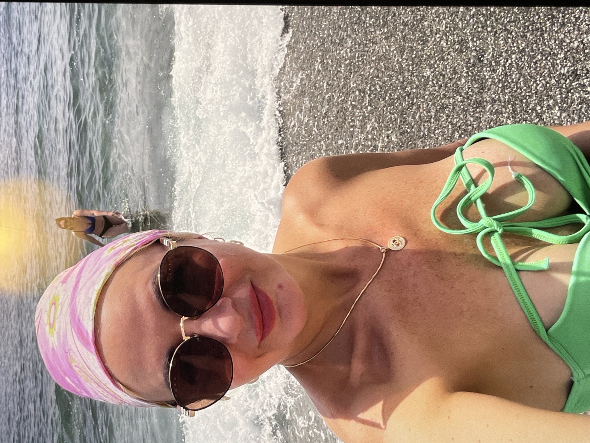 A woman wearing a pink headscarf and sunglasses is lying on a beach next to the ocean, wearing a green bikini and gold necklace. Another person is in the water in the background. The beach is comprised of grey sand.