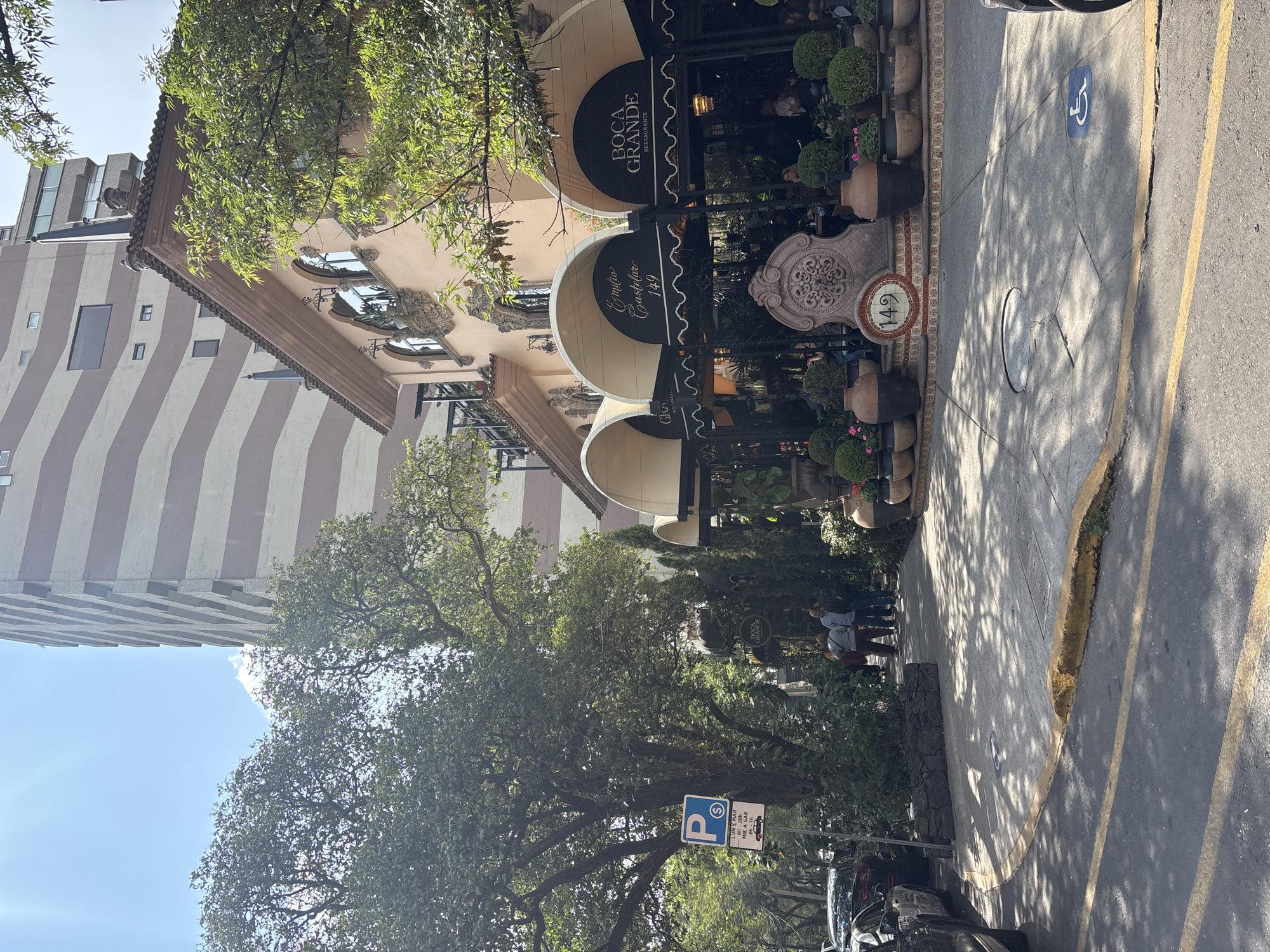 This image captures a vibrant daytime street scene in Mexico City, featuring a restaurant with outdoor seating. The main subject is a multi-story building with a light-colored, ornate facade, housing "BOCA GRANDE RESTAURANTE." Large dark awnings extend over the outdoor dining area, where several people are visible, some seated, others standing. The outdoor space is richly decorated with numerous potted plants, hedges, and flowers, alongside large urns and a decorative stone feature.
Text visible on the awnings includes "BOCA GRANDE RESTAURANTE" and "Emilio Castelar 149." The street address "149" is also displayed on a tiled sign on the sidewalk. The paved sidewalk shows distinct shadows from overhead trees and features a blue wheelchair accessibility symbol. An asphalt road with yellow markings and a curb runs alongside the sidewalk, with parked cars visible.
In the background, a tall, modern building with prominent vertical light and dark stripes rises against a clear blue sky. Lush green trees are scattered throughout the scene, casting shadows on the ground. A street sign indicates parking ("P" and "$") with specific hours: "LUN Y MAR 8h-20h" (Monday & Tuesday 8 AM - 8 PM) and "MIE A SAB 8h-1h" (Wednesday to Saturday 8 AM - 1 AM). The overall impression is a bustling, sunny day in a commercial or upscale neighborhood.