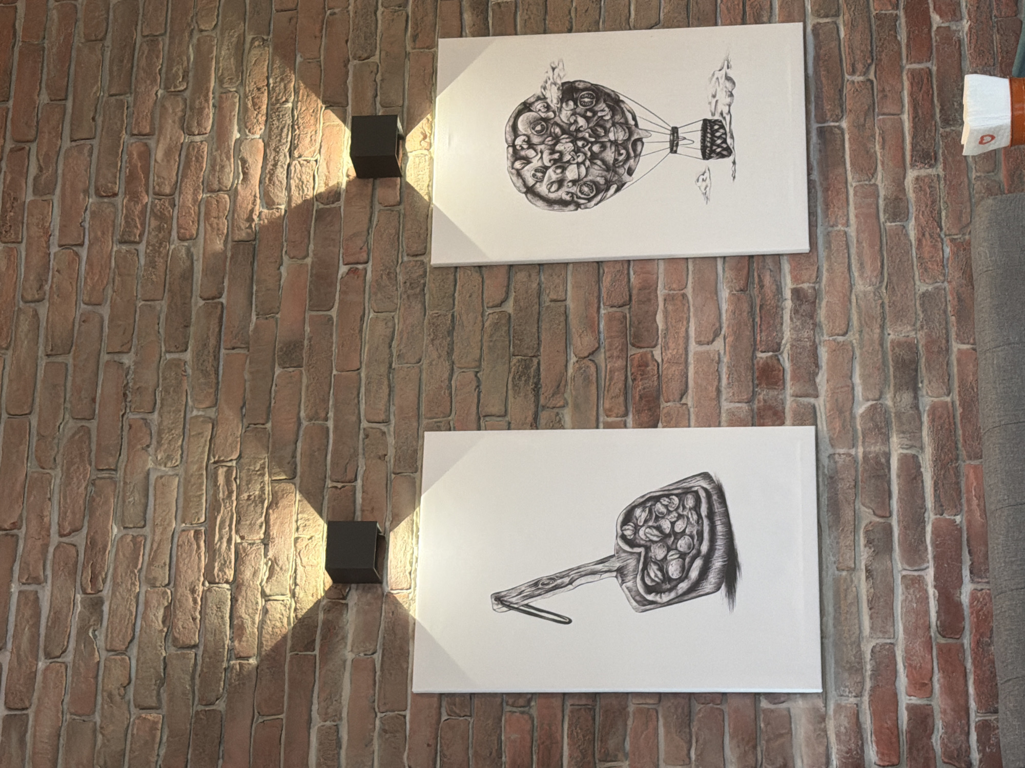 The image shows a close-up view of a brick wall indoors. Two black, cube-shaped light fixtures are mounted on the wall, one above the other, casting beams of light. Two white rectangular canvases hang on the wall between the lights. The top canvas displays a black-and-white drawing of a hot air balloon made of pizza. The bottom canvas features a black-and-white drawing of a pizza on a pizza peel. A portion of a gray object is visible on the right side of the frame. The location is most likely a pizzeria or restaurant in Burgas, Bulgaria.