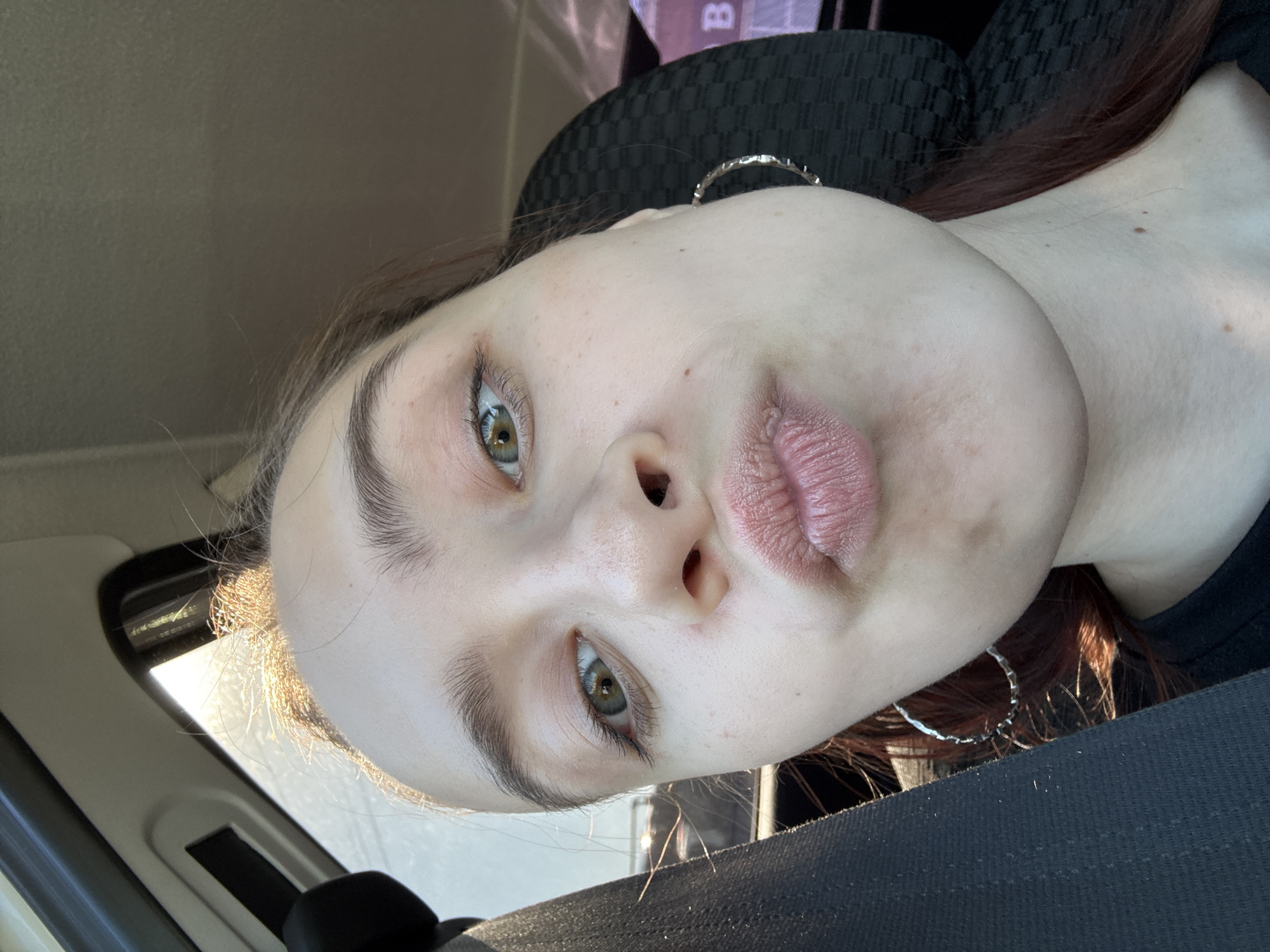 A close-up, rotated image captures the face of a person, likely a young female, situated within what appears to be the interior of a car. The person is looking directly at the camera with a playful expression, evidenced by puckered lips. Their eyes are open, revealing green or hazel irises with reflections of light, and they have distinct dark eyebrows and eyelashes. The skin appears fair, with visible freckles or small moles on the cheeks and forehead.
The setting is an automobile, indicated by a visible car seatbelt across the lower right, part of a car seat or headrest behind the head, and a car window frame on the left, letting in bright sunlight. The bright light suggests it is daytime and likely sunny, with some golden backlighting highlighting the hair around the temples. The person has dark reddish-brown hair, and large silver hoop earrings are visible on both sides of the head. In the blurry background, a faint letter "B" can be discerned on what seems to be part of the car's interior or an object outside the window. No other discernible text is visible.